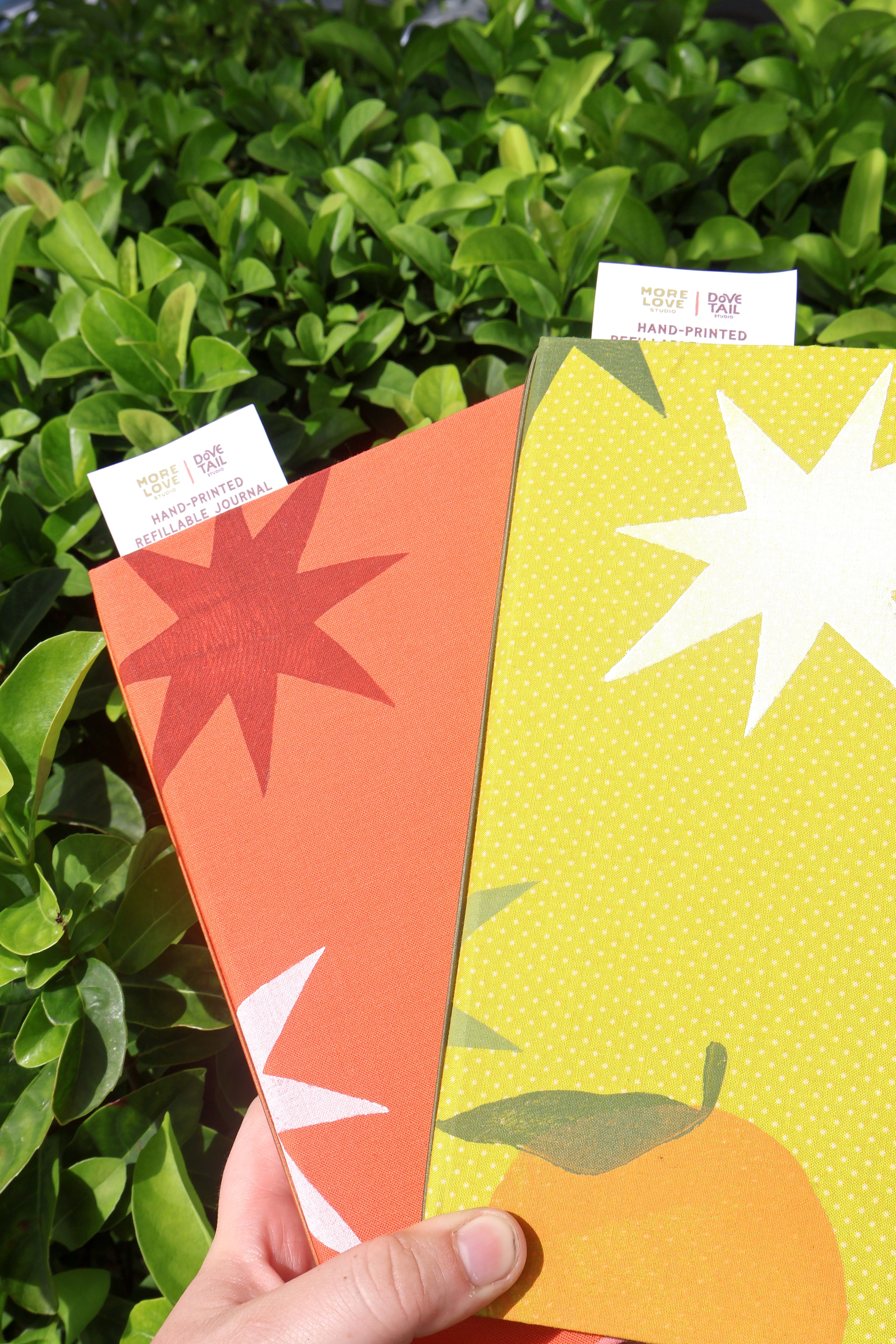 A hand holds two colorful patterned refillable bullet burnals in front of green foliage. One has an orange-red starburst on a coral background, and the other has a white starburst on a yellow dotted background.