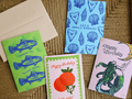 Four colorful greeting cards and an envelope arranged on a woven brown surface. The cards feature blue fish, seashells, a mermaid, oranges, and 'Happy Birthday' text in different designs.