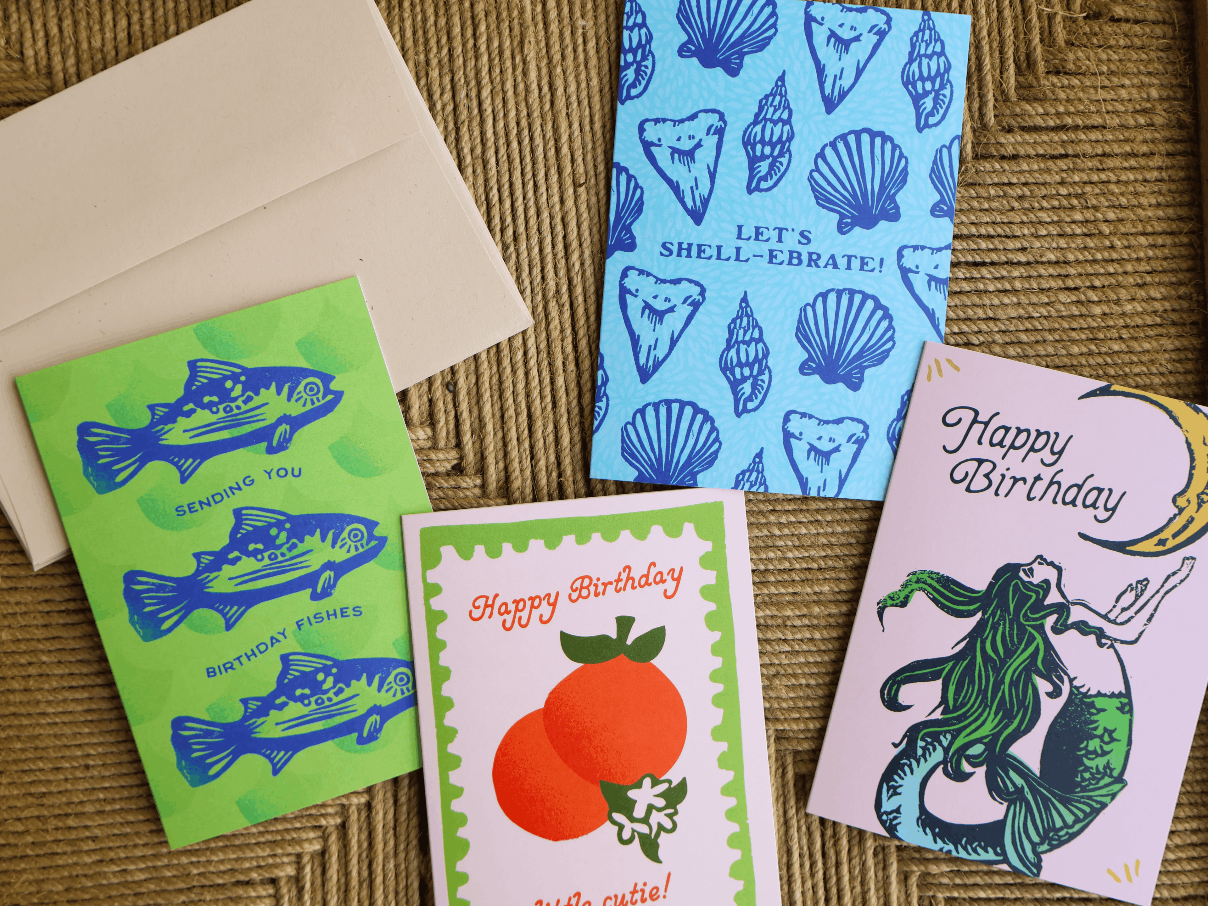 Four colorful greeting cards and an envelope arranged on a woven brown surface. The cards feature blue fish, seashells, a mermaid, oranges, and 'Happy Birthday' text in different designs.