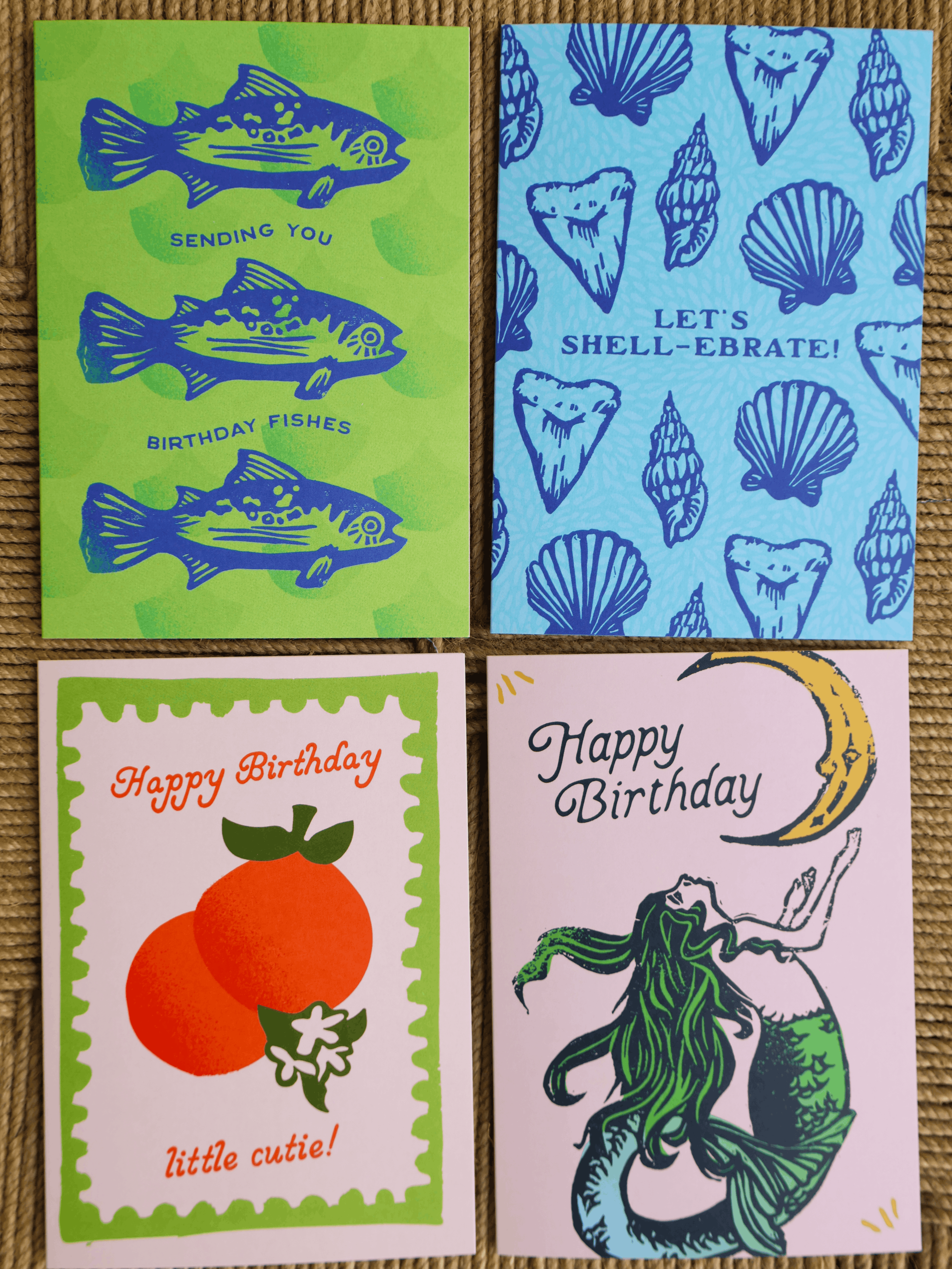 Four birthday greeting cards arranged in a 2x2 grid on a woven beige surface. The top-left card is lime green with blue fish illustrations and the text 'SENDING YOU BIRTHDAY FISHES.