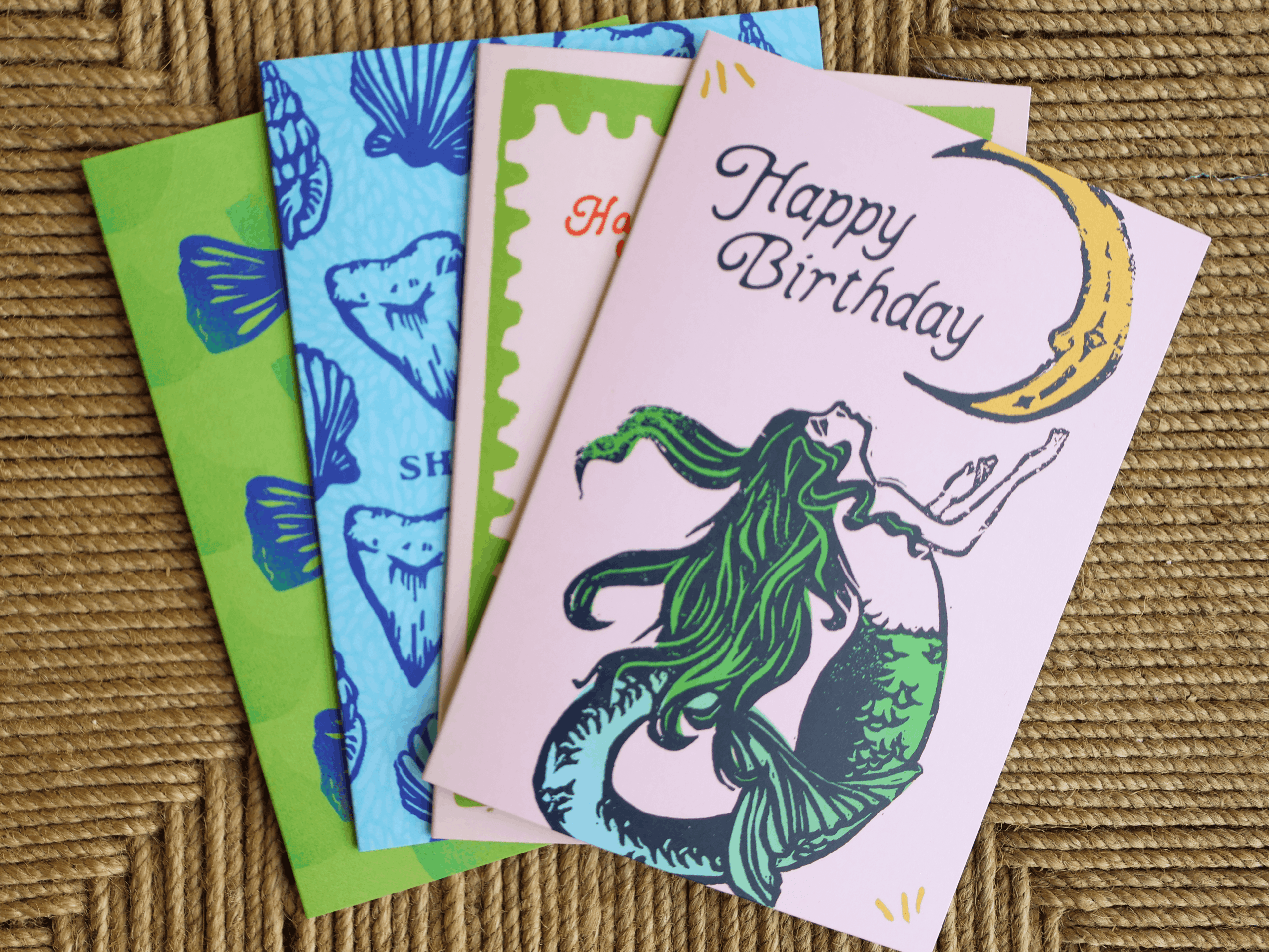 A set of colorful greeting cards fanned out on a woven brown surface. The front card is light pink with a mermaid illustration and the words 'Happy Birthday' in black script, alongside a yellow crescent moon.
