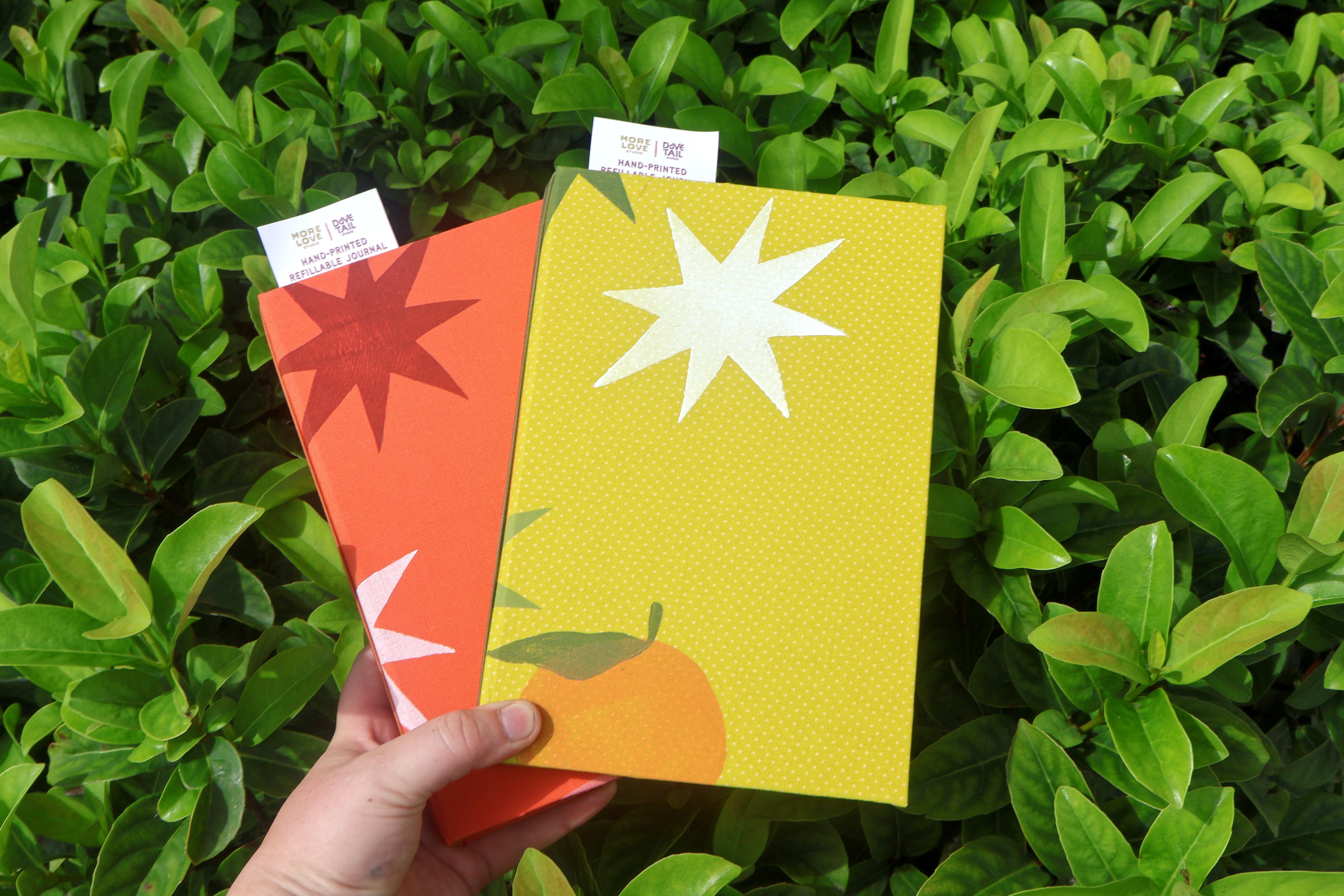A hand holds two colorful refillable journals in front of dense green foliage. The front journal is yellow with a large white starburst and a small orange fruit illustration, and the journal behind it is orange with a red starburst and white star accent. hand bound band linocut printed by more love studio
