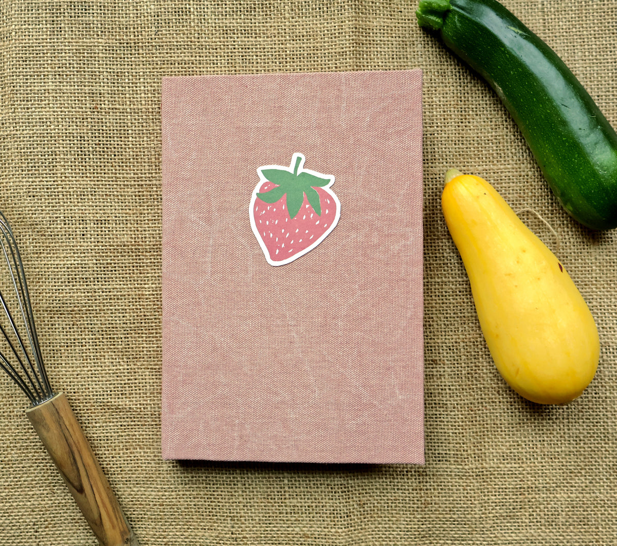 A pink textured hardcover recipe log with a strawberry linocut print design on the cover, shown next to an open matching book on a burlap background with a green cucumber and a yellow squash as props.