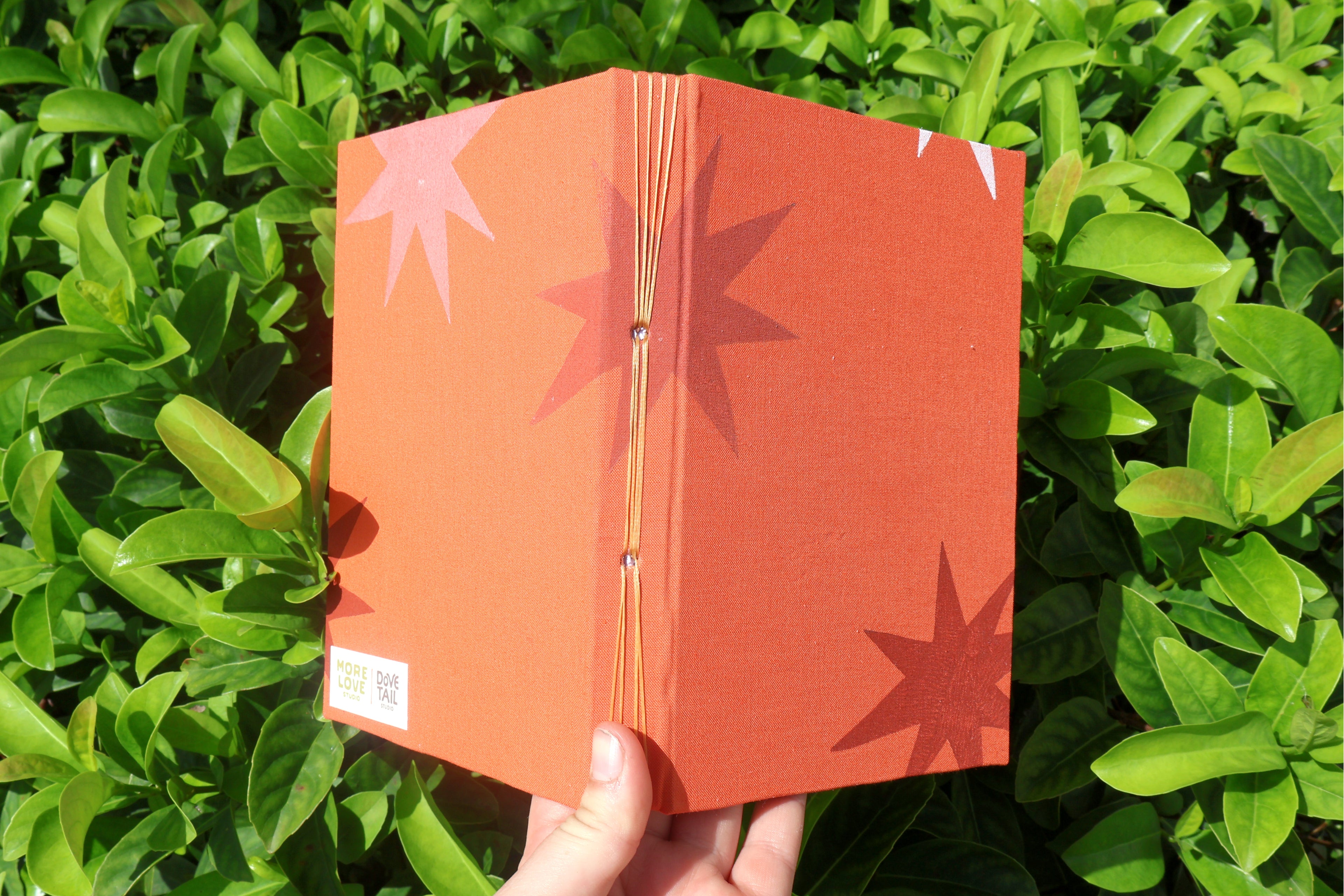 A hand holds a small orange sketchbook with star-shaped cutout designs, set against a background of green leaves. Hand-bound and linocut printed 