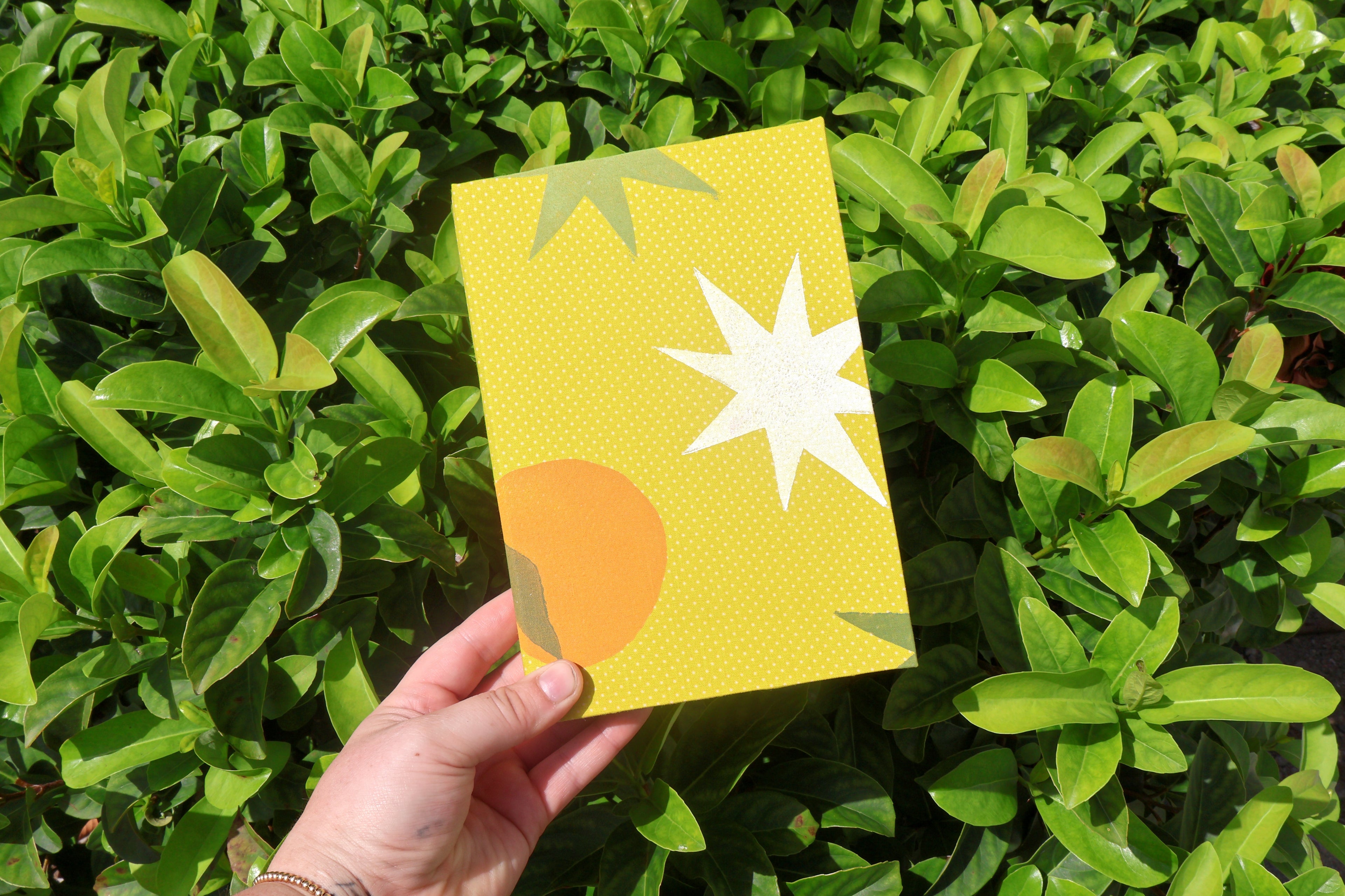 A hand holds a yellow watercolor sketchbook cover in front of dense green foliage. The cover has a yellow dotted texture and abstract geometric shapes, including a white starburst, an orange semicircle, and green leaf motifs. hand bound and linocut printed with more love studio