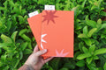A hand holds two orange refillable journals with minimalist starburst designs against a background of lush green leaves. One notebook features a dark red star, and the other features a white star. hand bound and linocut printed with more love studio
