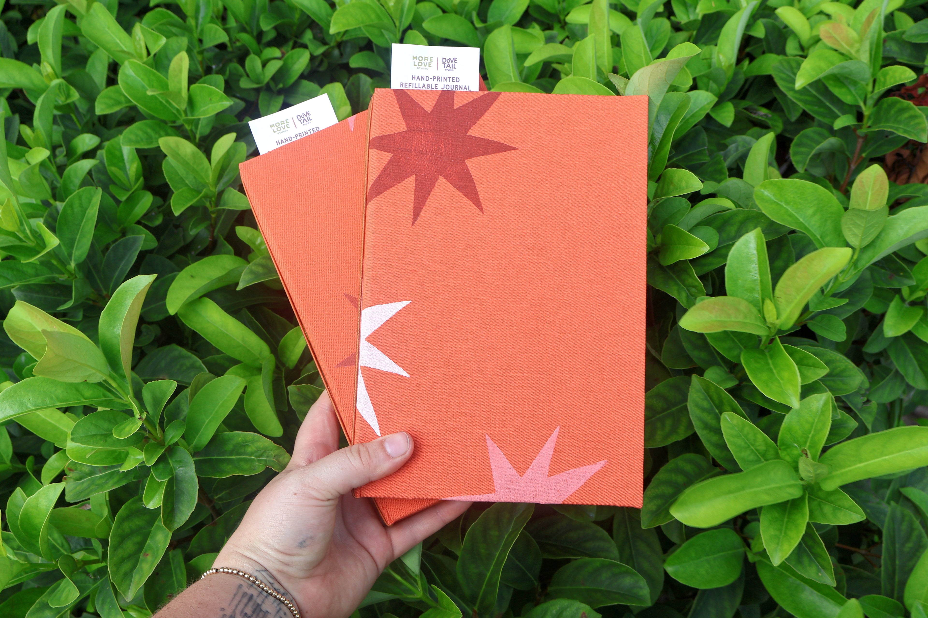 A hand holds two orange refillable journals with minimalist starburst designs against a background of lush green leaves. One notebook features a dark red star, and the other features a white star. hand bound and linocut printed with more love studio