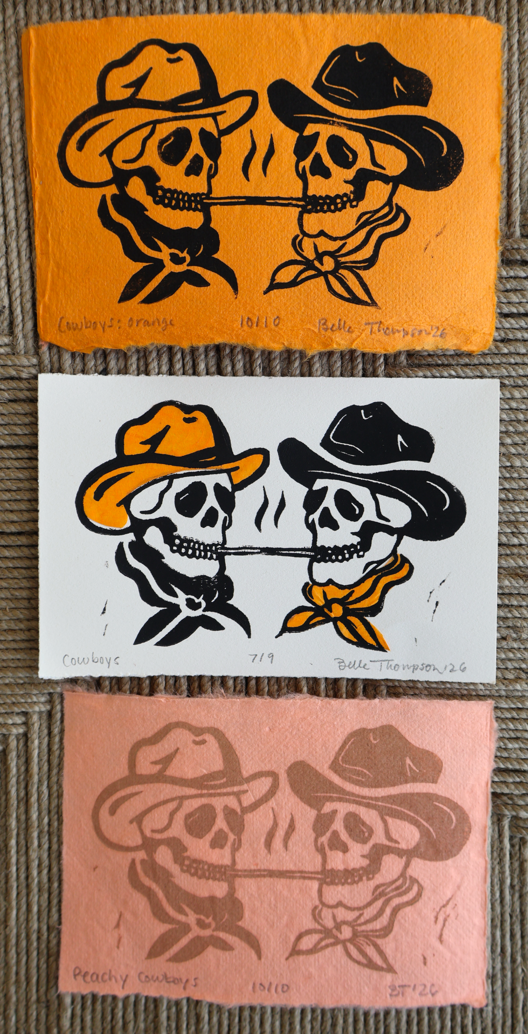 Three hand-colored art prints depicting two cowboy skulls smoking cigars, shown stacked vertically on a woven mat. The prints feature orange, white, and dusty pink backgrounds with black ink illustrations.