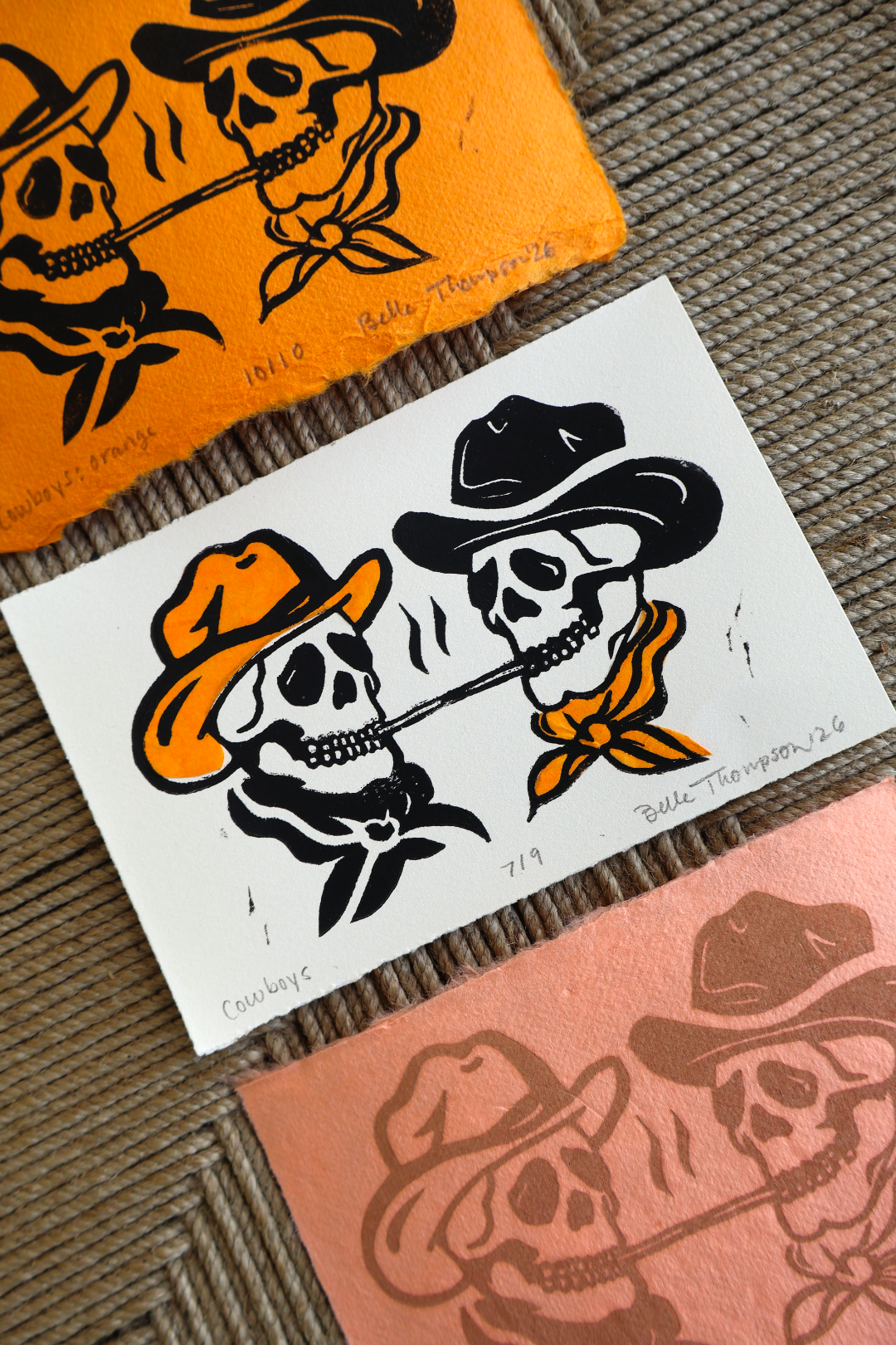 Three hand-colored art prints featuring a cowboy skull design, arranged diagonally on a textured brown woven mat. The prints use orange, white, and dusty rose paper with black ink, and are signed by Bella Thompson.