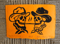 An orange rectangular art print or card featuring a black ink illustration of two cowboy skulls wearing hats and bandanas, facing each other with a line between them. Handwritten text appears along the bottom edge.
