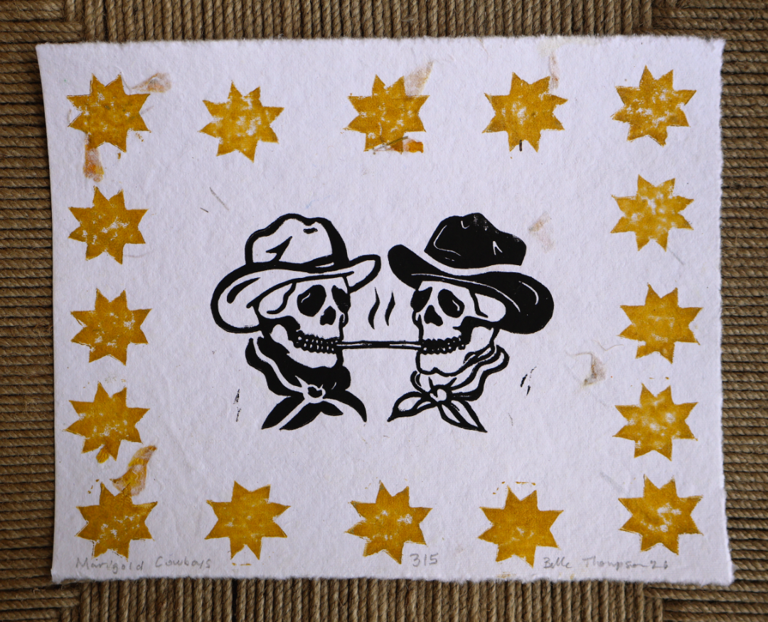 A square white art print or card featuring a western-themed design: two black-and-white skulls wearing hats facing each other, surrounded by a border of yellow starbursts, photographed flat against a brown woven surface.