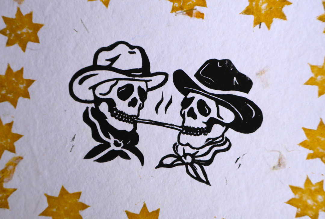 A close-up of a printed design featuring two black-and-white cowboy skulls wearing hats and bandanas, with scattered yellow star-like motifs on a white textured background.