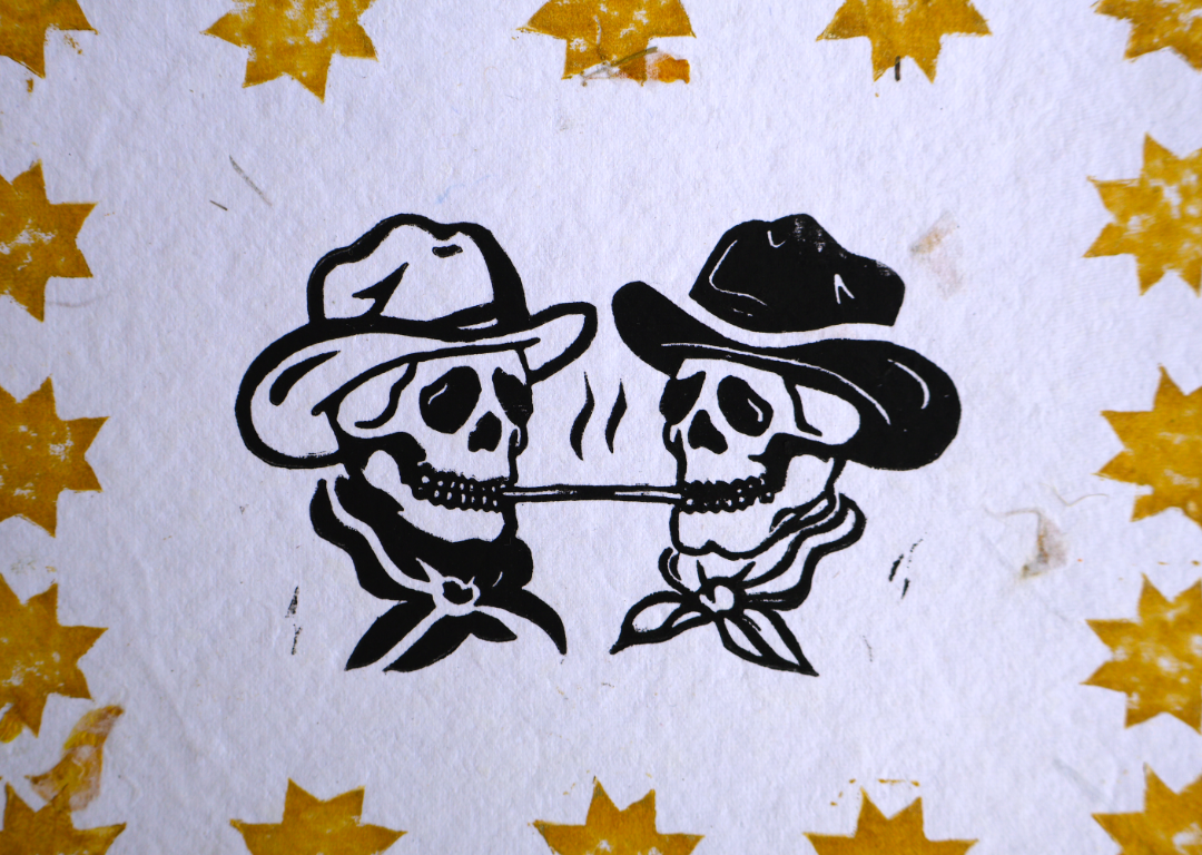 A close-up of a printed design featuring two black-and-white cowboy skulls wearing hats and bandanas, centered on a white textured background with scattered gold star motifs around the edges.