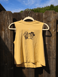 A sleeveless yellow t-shirt with a black graphic of two cartoon figures wearing cowboy hats, hanging on a wooden hanger against a weathered brown wooden fence with blue sky and trees in the background.