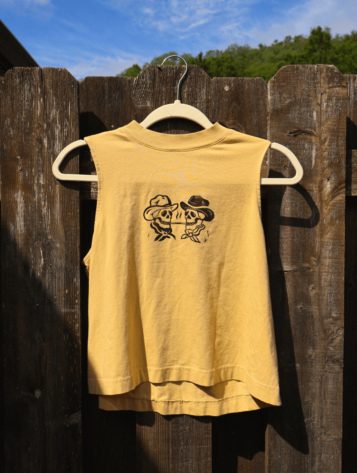 A sleeveless yellow t-shirt with a black graphic of two cartoon figures wearing cowboy hats, hanging on a wooden hanger against a weathered brown wooden fence with blue sky and trees in the background.
