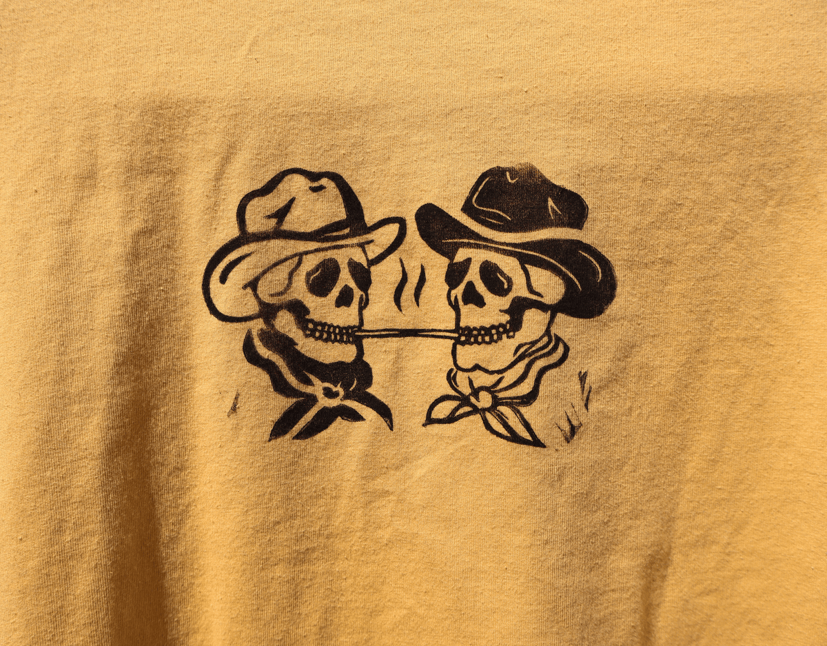 A person's hands hold up a yellow sleeveless tank top with a black graphic of two cartoon figures wearing cowboy hats, set against a dark, textured stone wall.