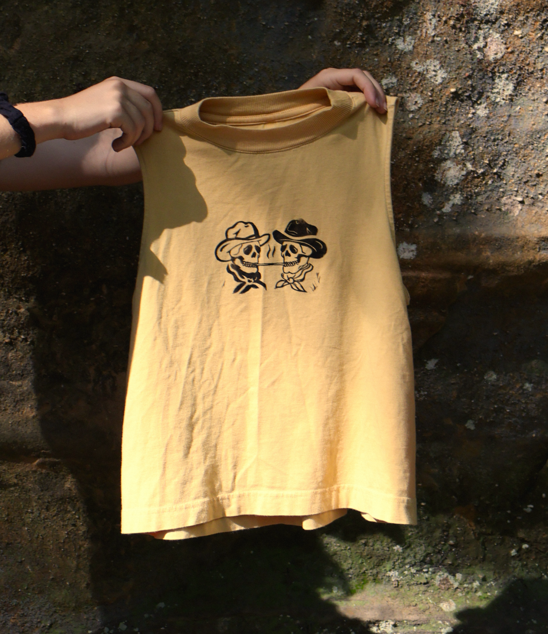 A person's hands hold up a yellow sleeveless tank top with a black graphic of two cartoon figures wearing cowboy hats, set against a dark, textured stone wall.