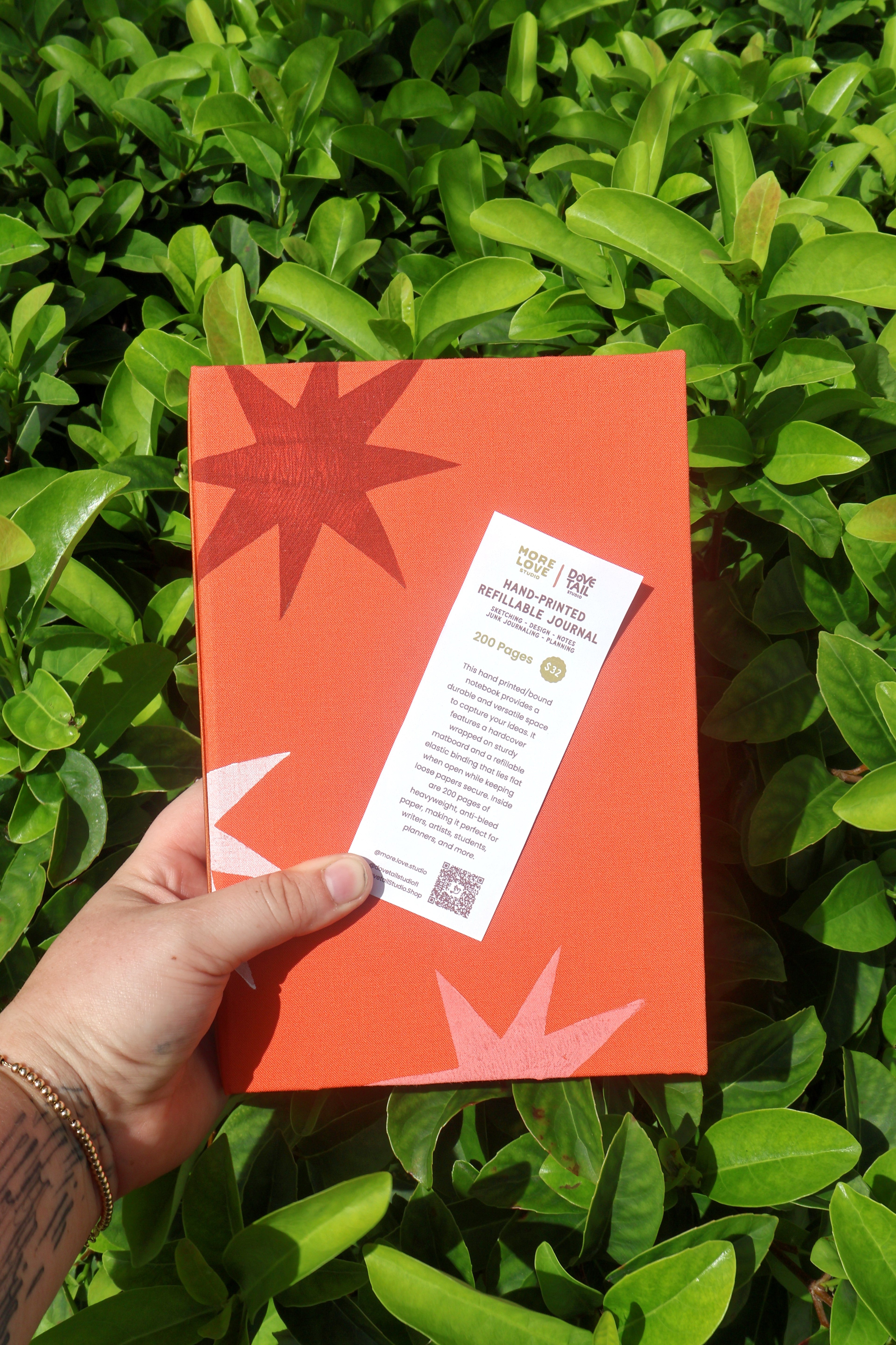 A hand holds a bright orange refillable journal against a background of lush green tea plant leaves. The cover features a bold red starburst and white star accents, with a white product information card attached. hand bound and linocut printed with more love studio