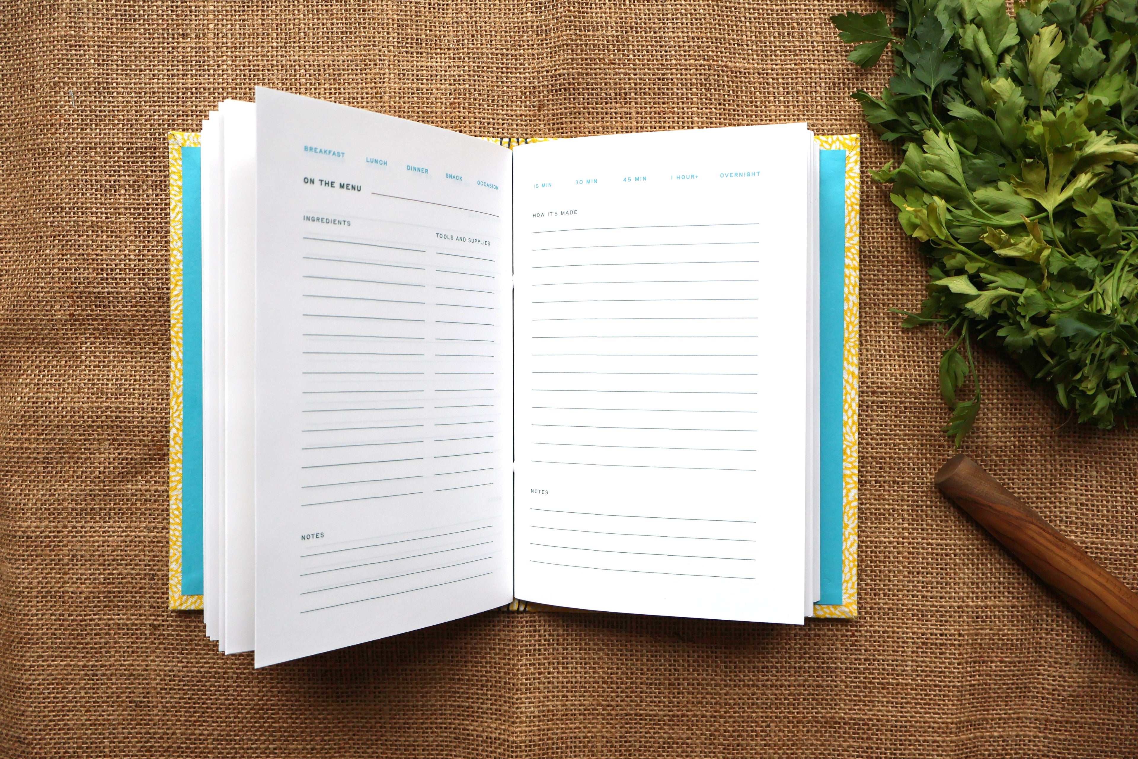 An open recipe log with white designed pages, blue headers, and a yellow-and-blue patterned cover, styled on a burlap surface with parsley and a wooden spoon nearby.