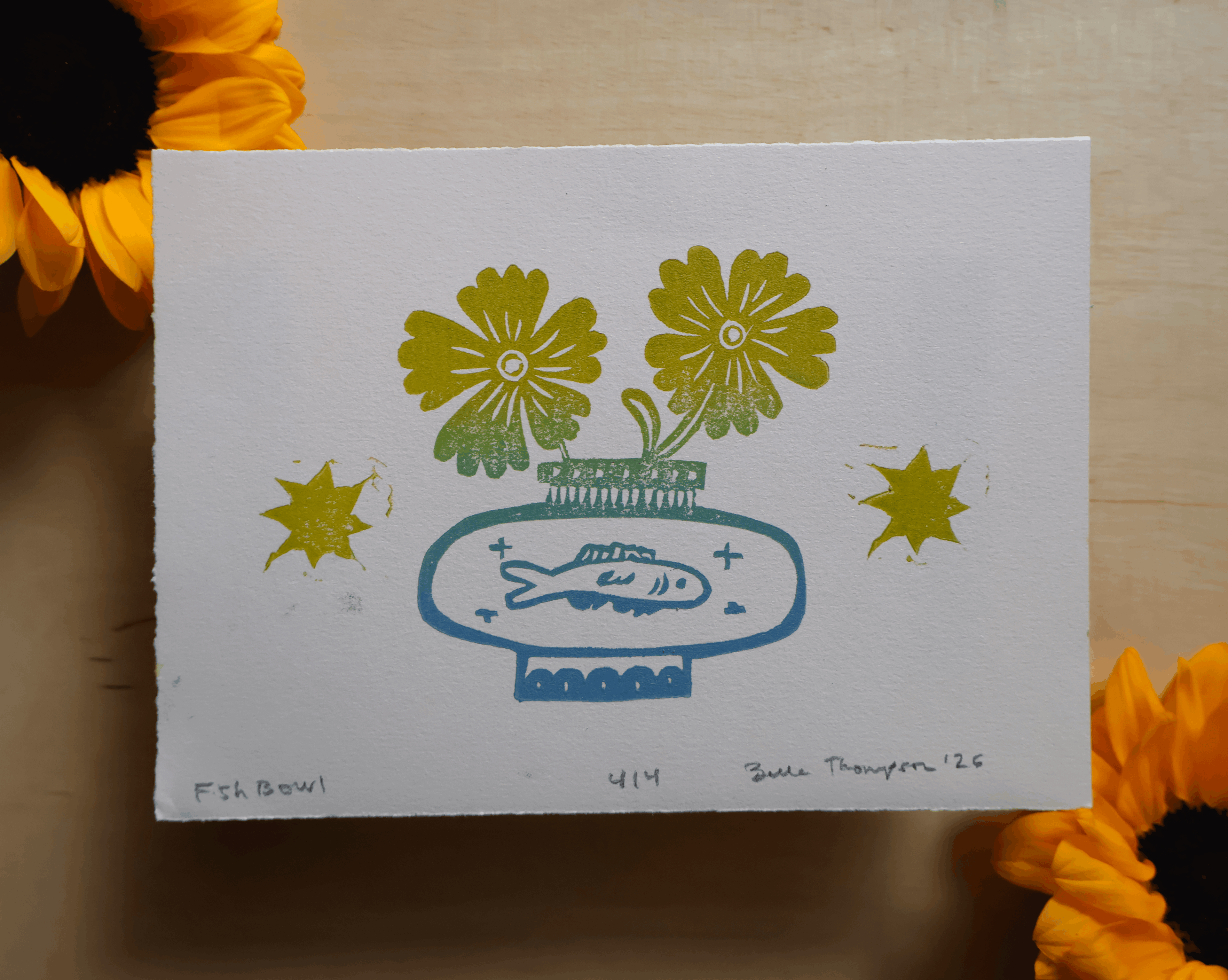 A square white art print or card featuring a stylized blue vase with a fish and two yellow flowers, placed on a light wood surface with sunflower petals around it. Signed and dated