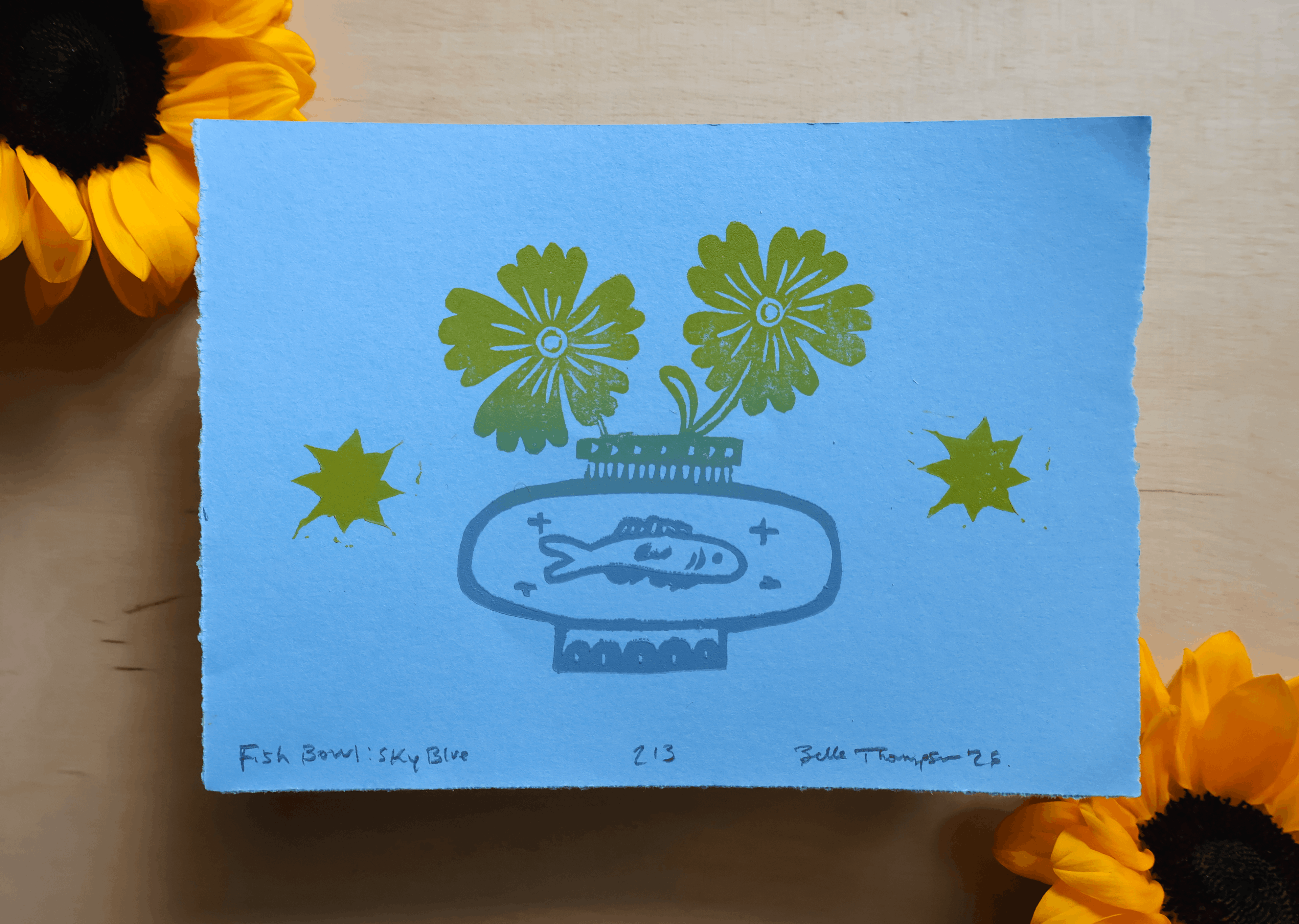 A square art print or card with a light blue background and a stylized green fish bowl illustration, placed on a light wood surface with yellow flowers around the edges.