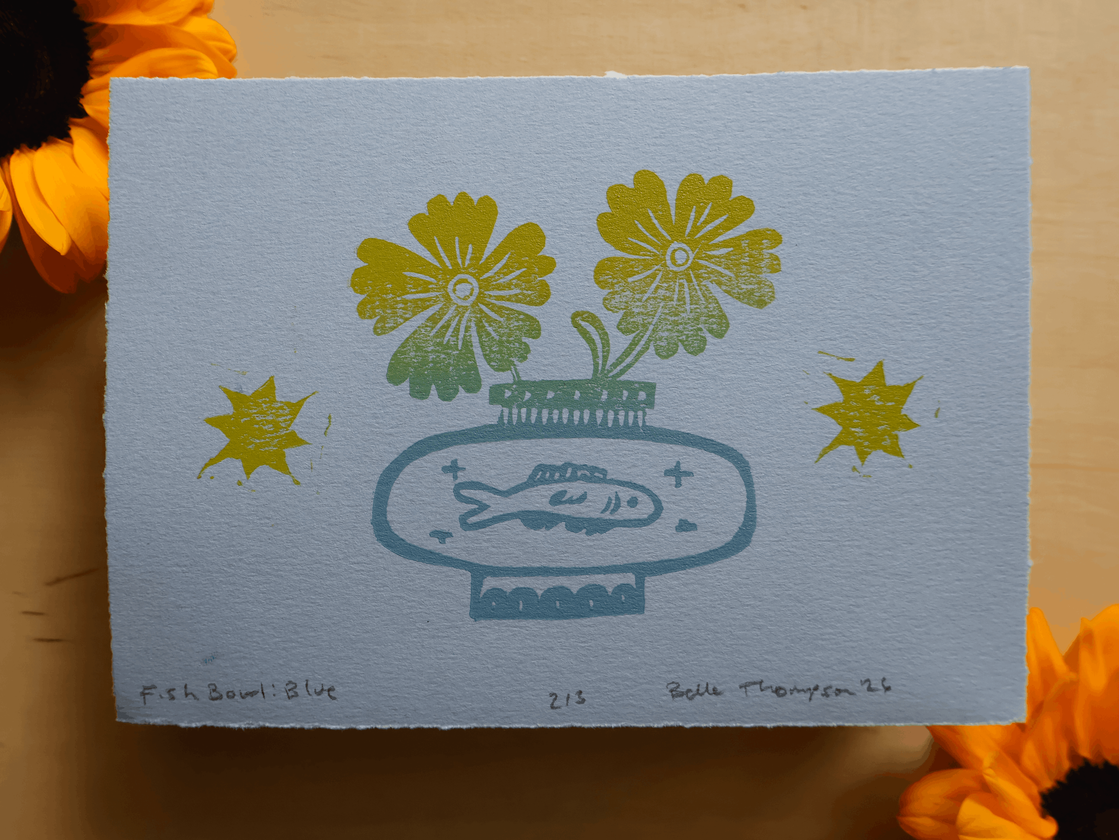 A square art print on textured paper featuring a blue fish in a vase with yellow flowers, styled with orange flowers in the corners on a wooden surface.