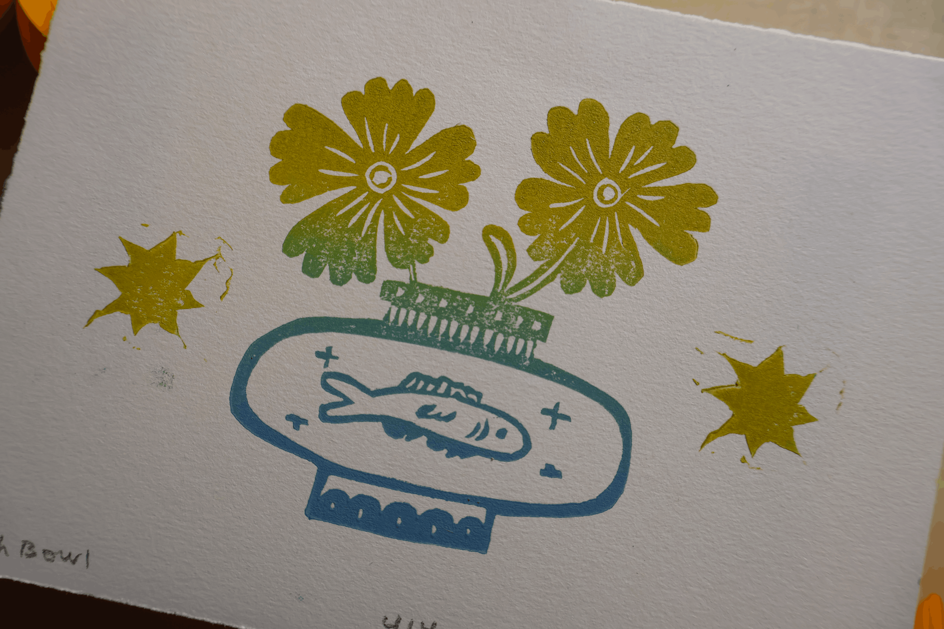 A close-up of a printed artwork on textured white paper, featuring two yellow flowers with green stems in a blue bowl with a fish illustration, plus yellow star-like shapes on either side.