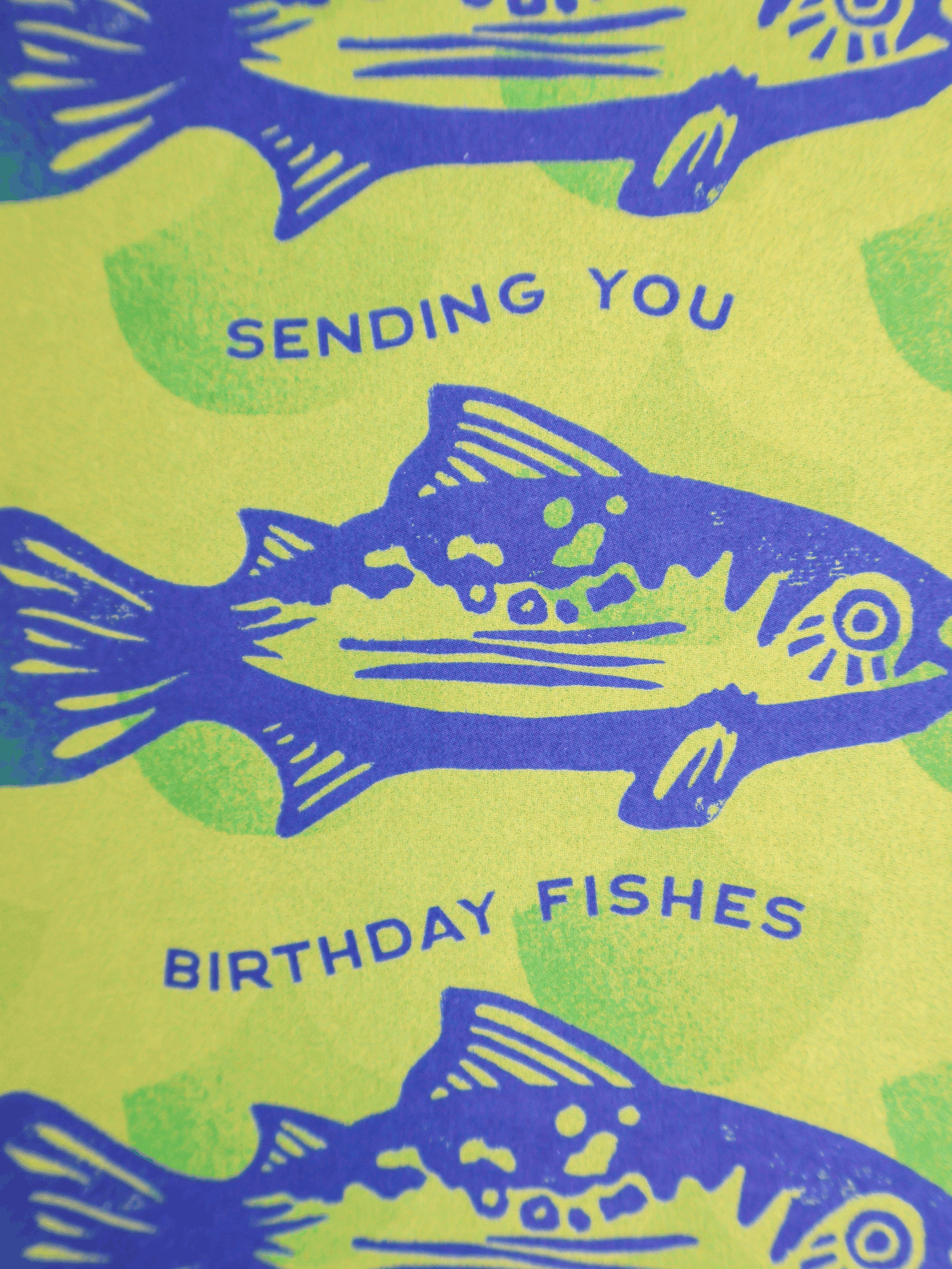 A close-up of a printed card or paper design featuring blue fish illustrations and the text 'SENDING YOU BIRTHDAY FISHES' on a bright yellow-green background with pale green splotches.