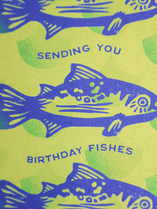 A close-up of a printed card or paper design featuring blue fish illustrations and the text 'SENDING YOU BIRTHDAY FISHES' on a bright yellow-green background with pale green splotches.