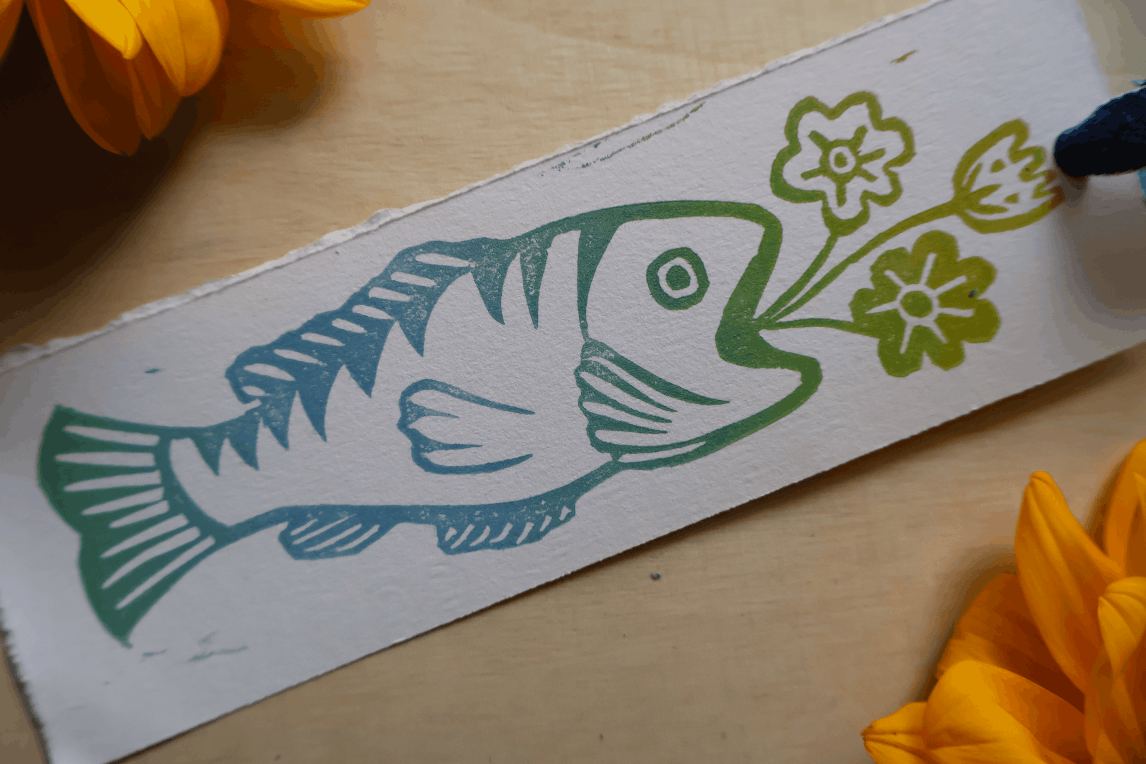 A handmade paper art print featuring a stylized fish and flowers in green and blue-green ink, shown on a light wood surface with yellow flowers in the corners.