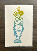A square art print on textured off-white paper featuring a stylized fish in teal and green with yellow flowers on a dark wood background. Handwritten text appears at the bottom of the print.