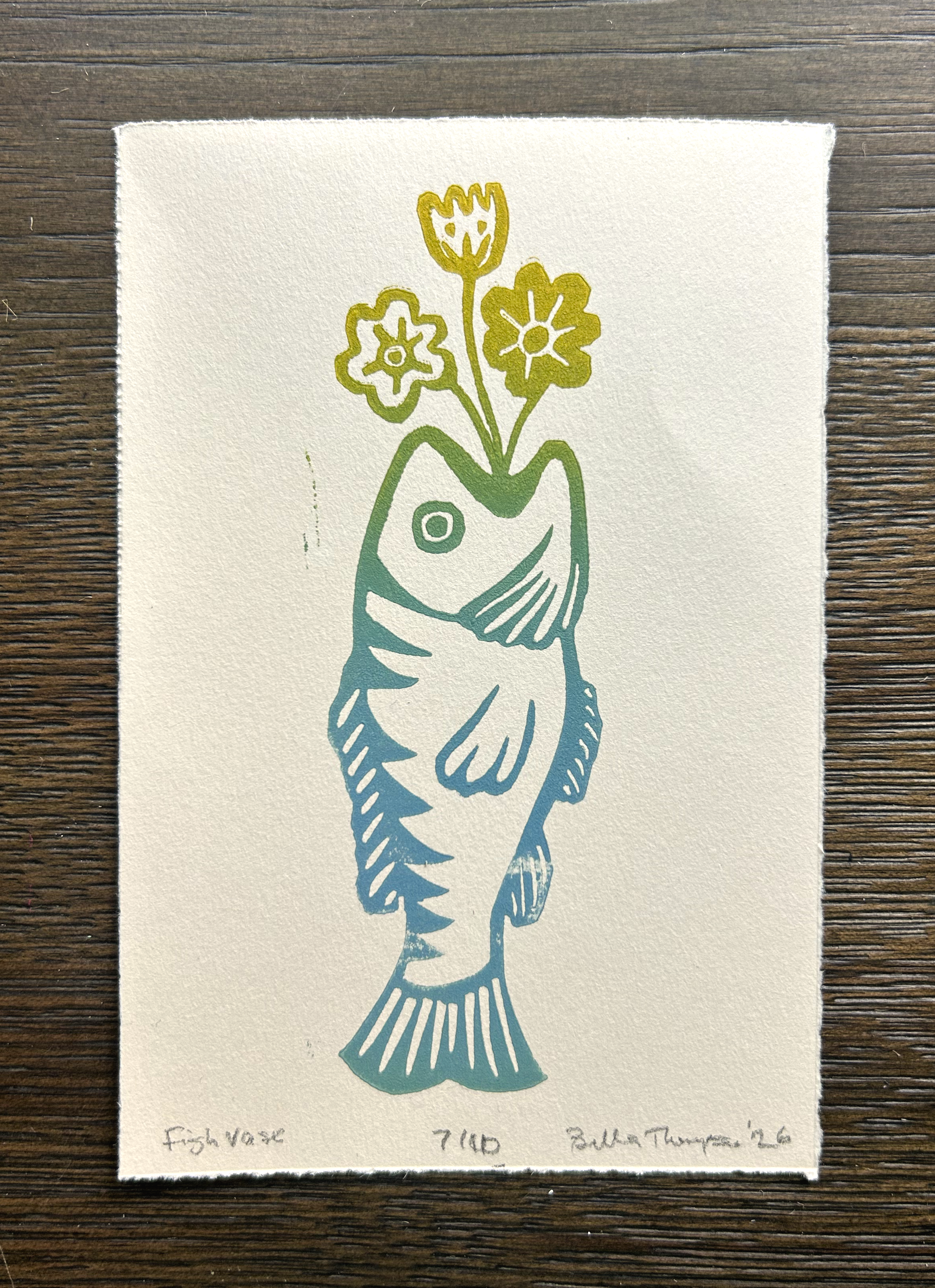 A square art print on textured off-white paper featuring a stylized fish in teal and green with yellow flowers on a dark wood background. Handwritten text appears at the bottom of the print.