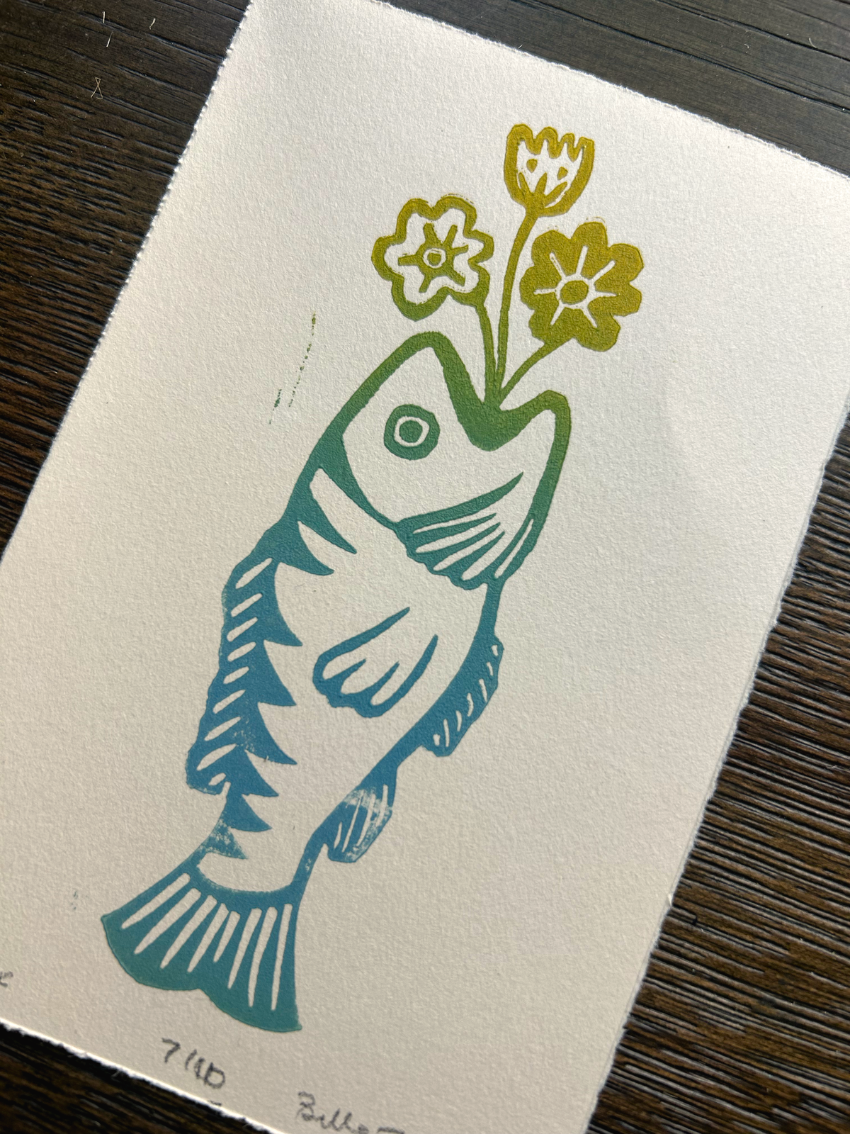 A square art print on textured off-white paper featuring a stylized fish in teal and green with yellow flowers in its mouth, placed on a dark wood surface.