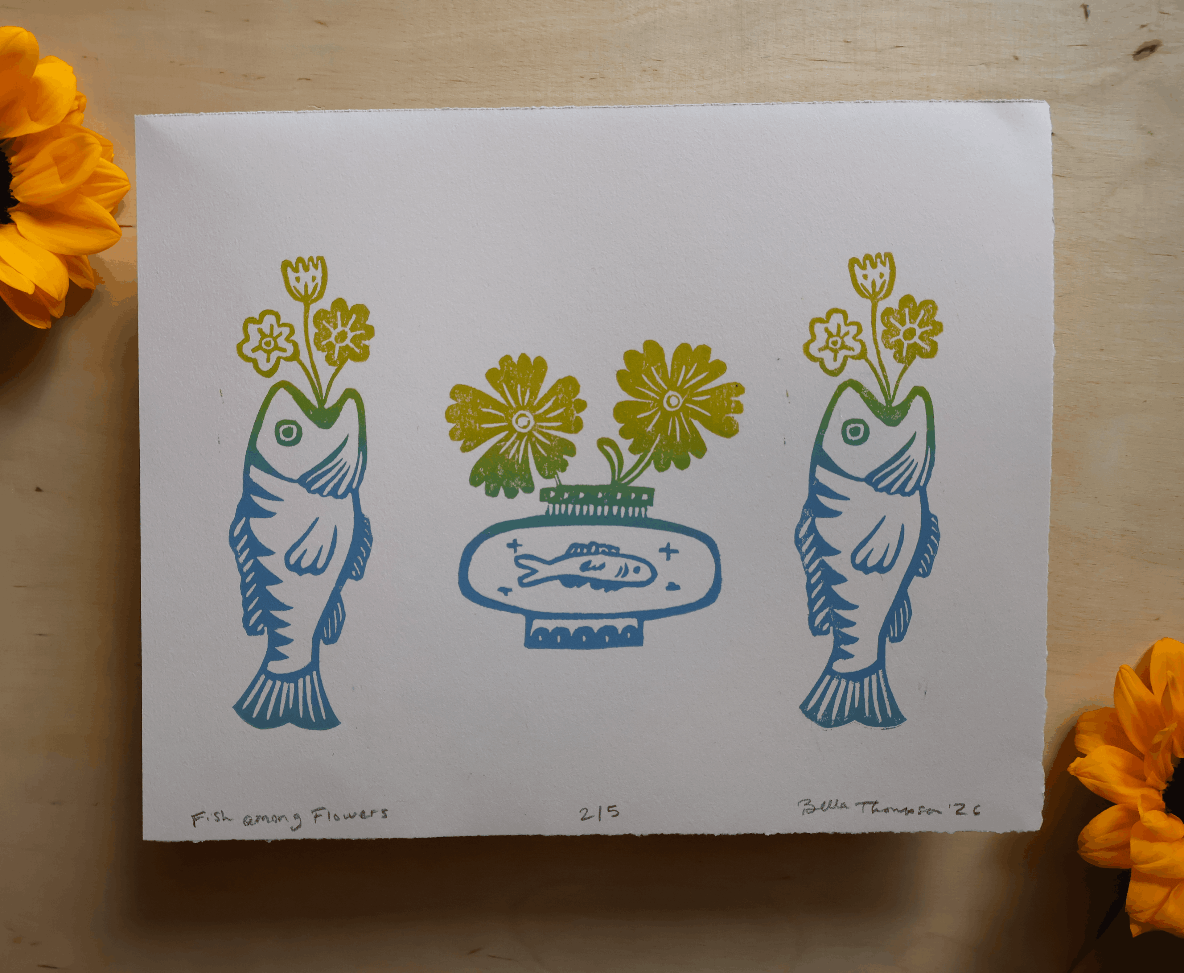 A square white art print featuring a minimalist blue and yellow linocut-style design of two fish holding flower bouquets and a central vase with flowers, photographed on a light wood surface with yellow flowers at the corners.