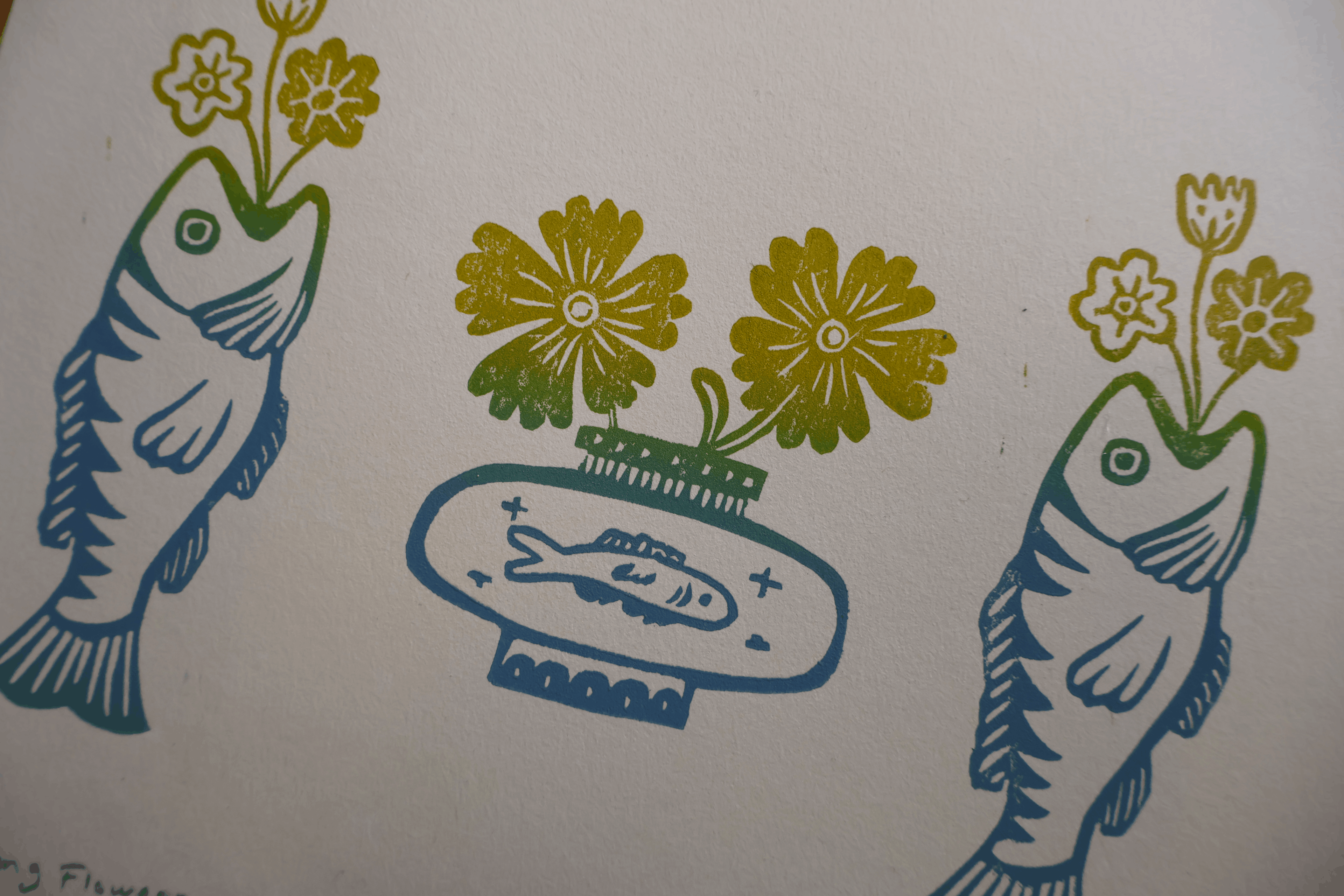A close-up of a printed or painted design on an off-white background, showing two stylized fish in blue-green outlines with yellow flowers above them, and a central fish-shaped vase with yellow flowers.