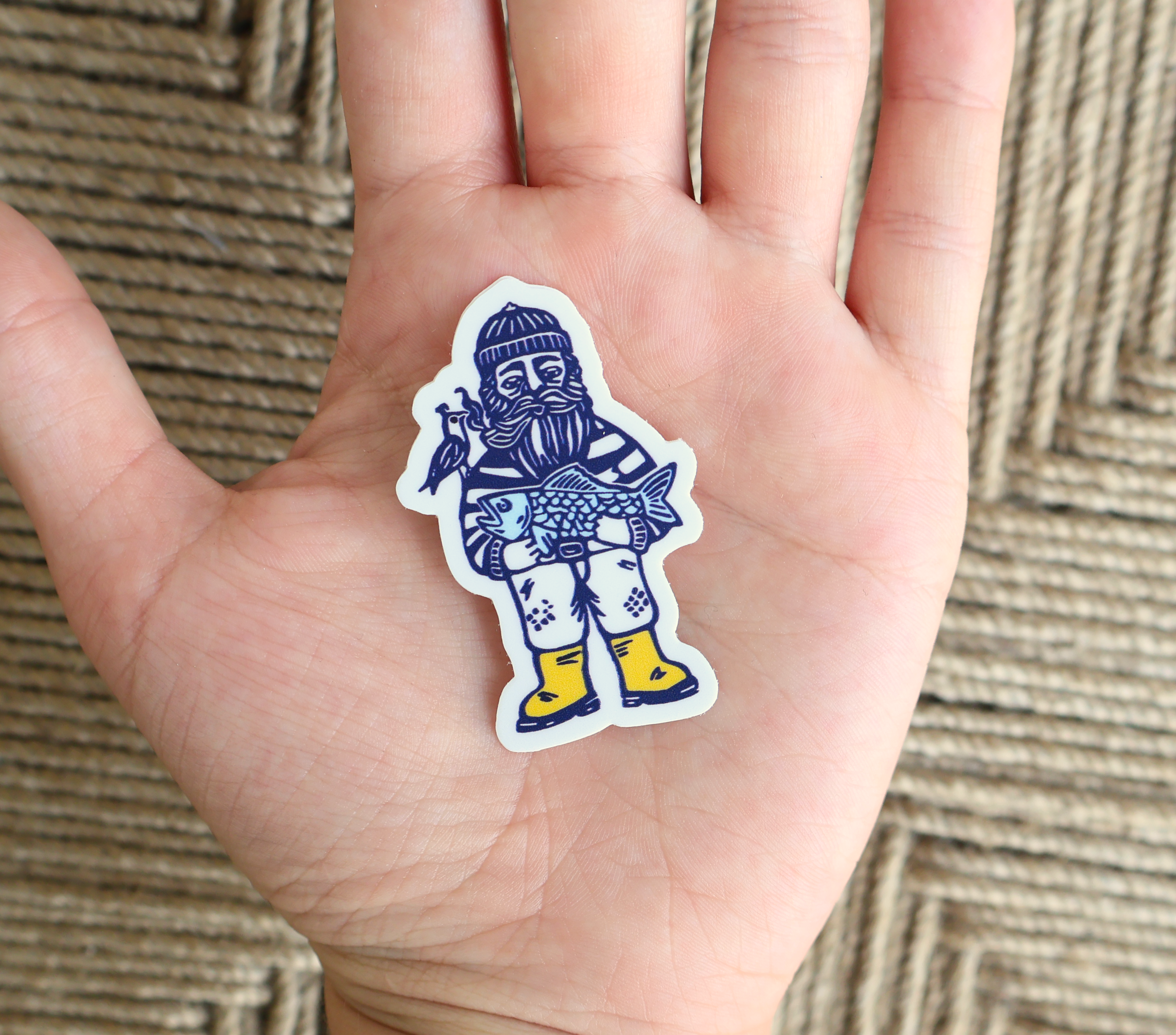 A die-cut sticker featuring a stylized bearded man in winter clothing holding a fish, placed on an open hand. The sticker has a white border and is shown against a woven beige background.