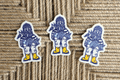 Three die-cut stickers featuring a stylized blue and white fisherman holding a fish, with yellow boots, arranged on a woven jute background.