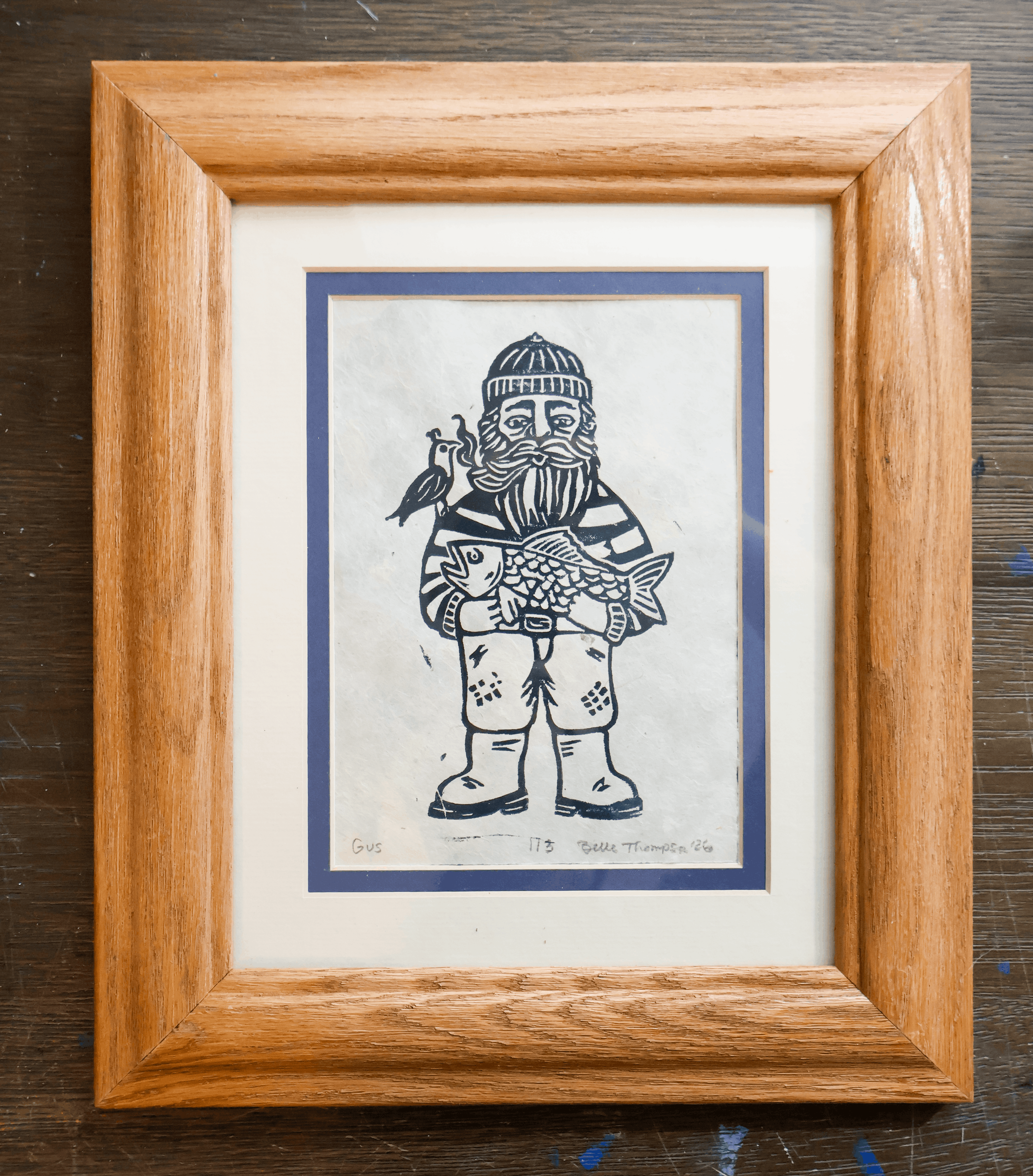 A framed art print featuring a black and white woodcut-style illustration of a bearded fisherman holding a fish, surrounded by a white mat and a natural wood frame, set against a dark wooden tabletop.