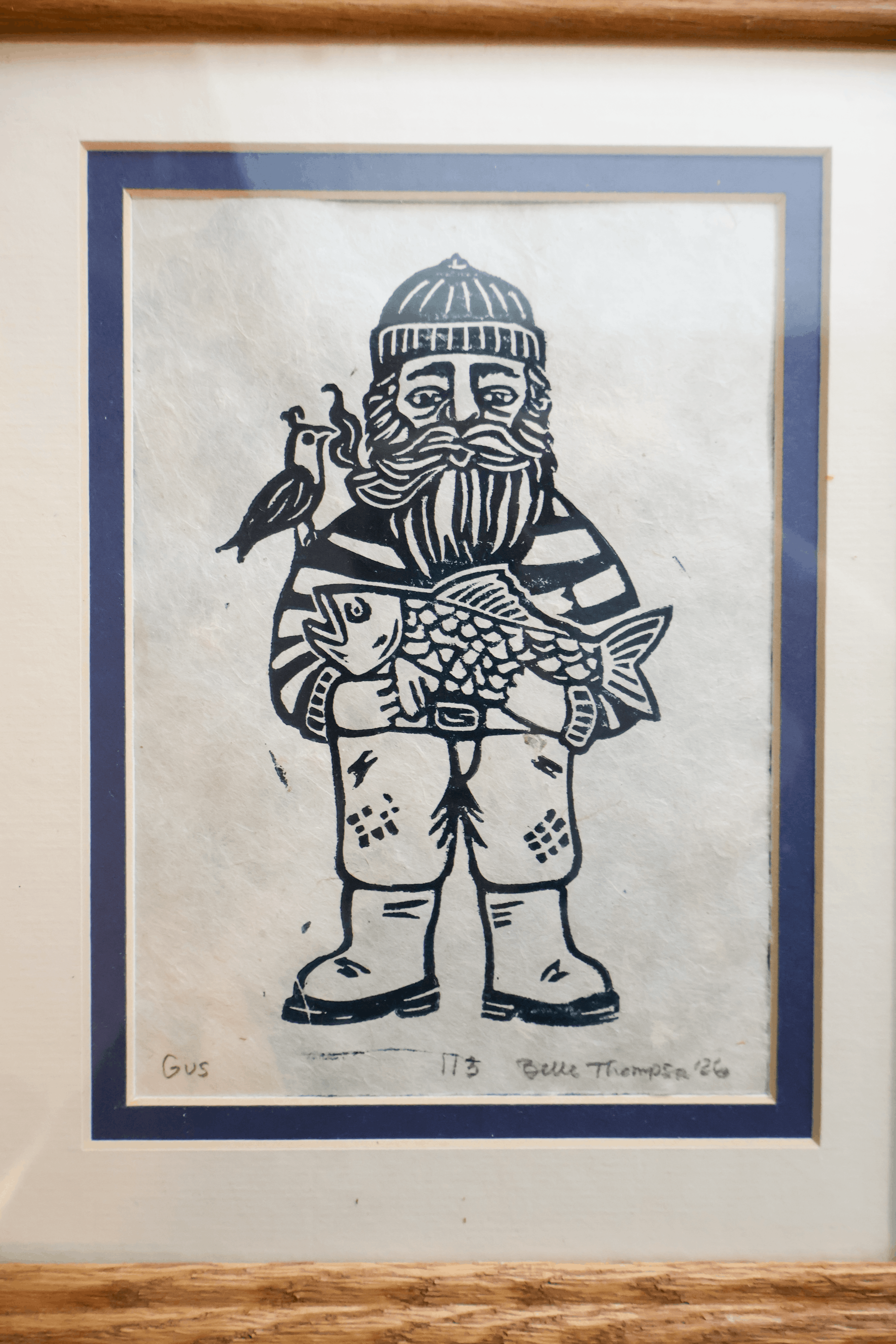 A framed art print featuring a black and white woodcut-style illustration of a bearded man in winter clothing holding a fish, with a bird on his shoulder. The print is mounted with a blue mat border and a light-colored mat, set within a natural wood frame.