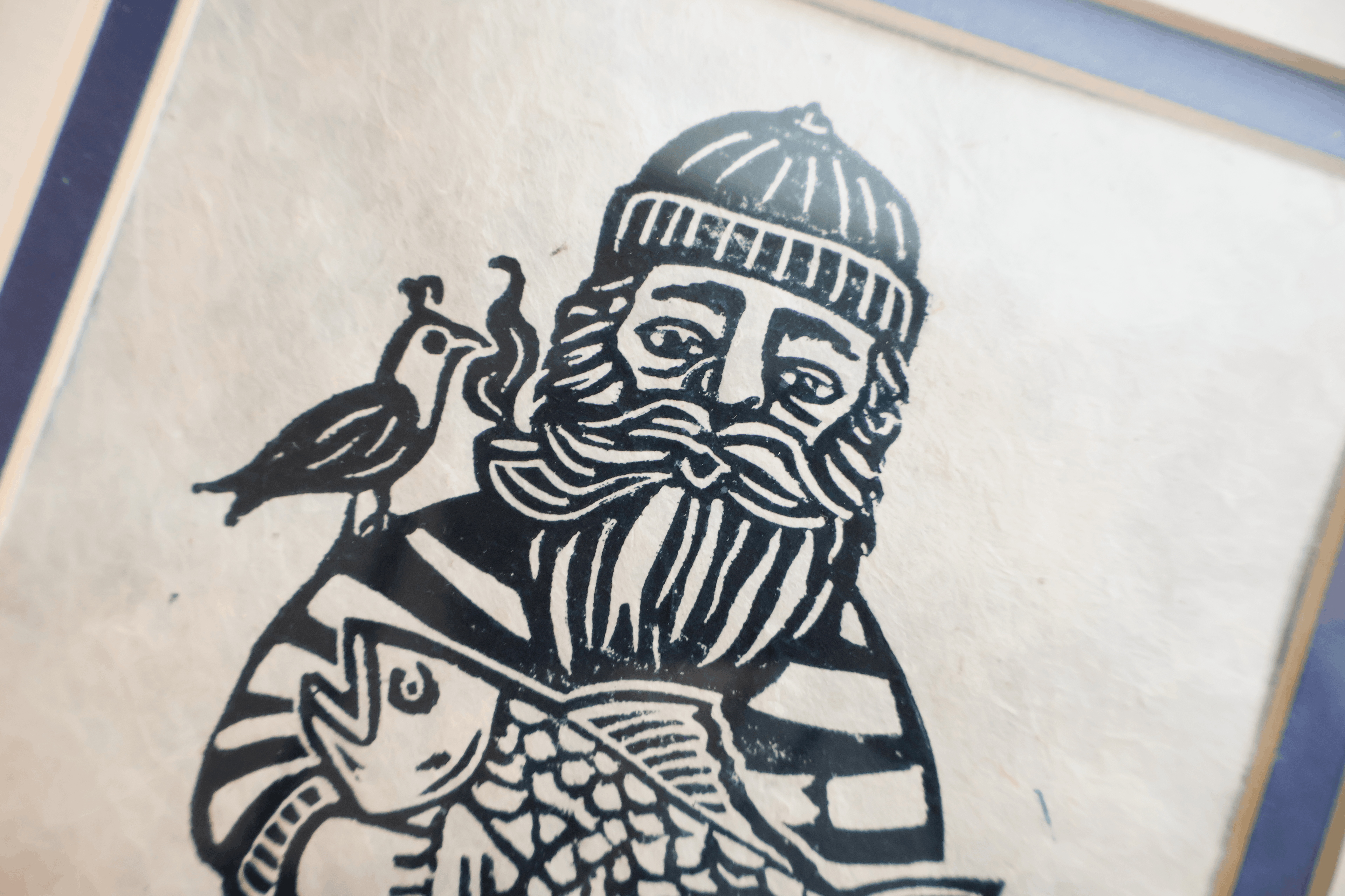 A close-up of a black ink block print or woodcut-style artwork on textured off-white paper, showing a bearded figure wearing a striped hat and holding a fish, with a bird on the shoulder. The print is bordered by blue and gold-toned edges.