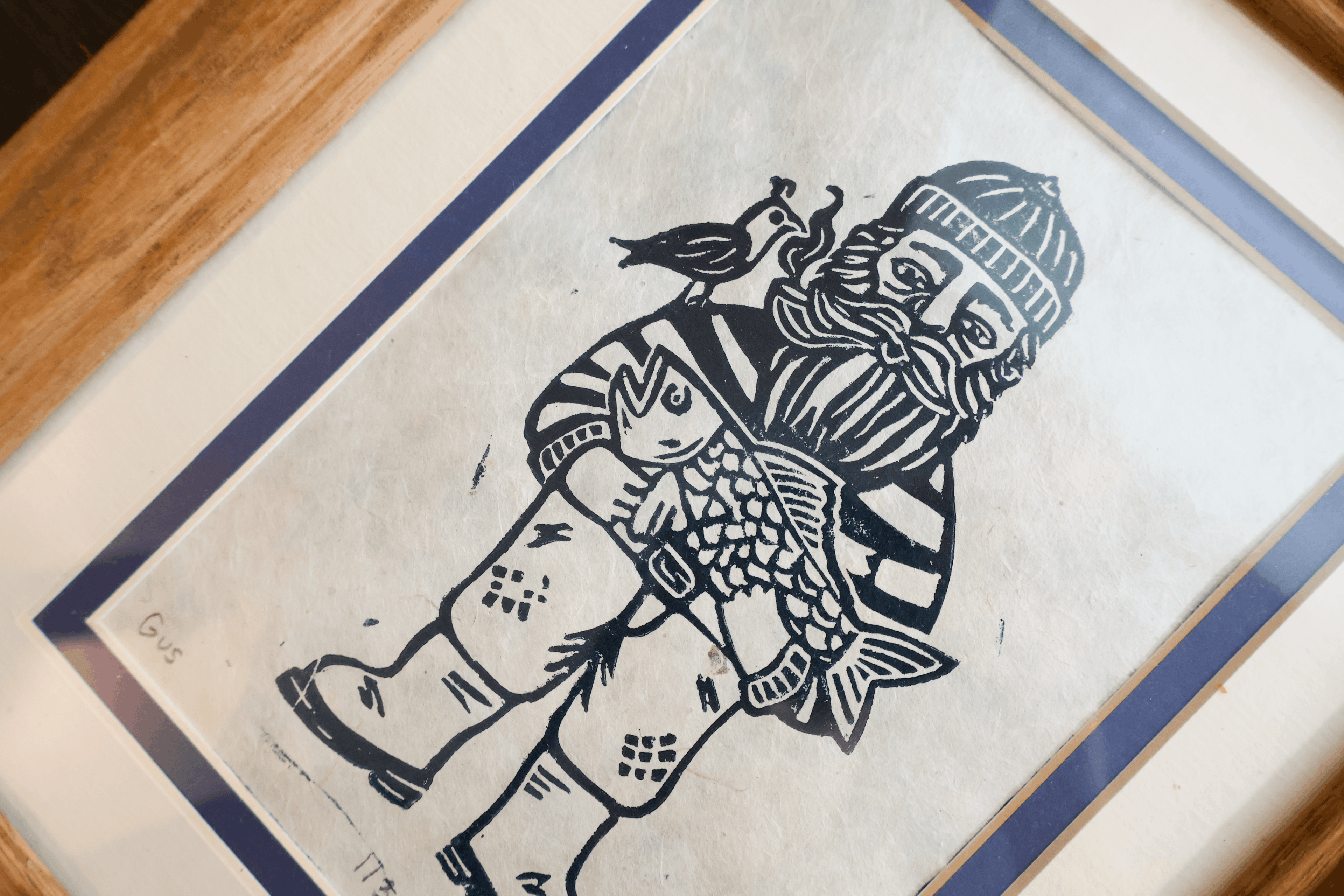 A framed black and white woodcut-style print showing a bearded figure in winter clothing holding a fish, with a bird on his shoulder, photographed at an angle over a wooden surface.