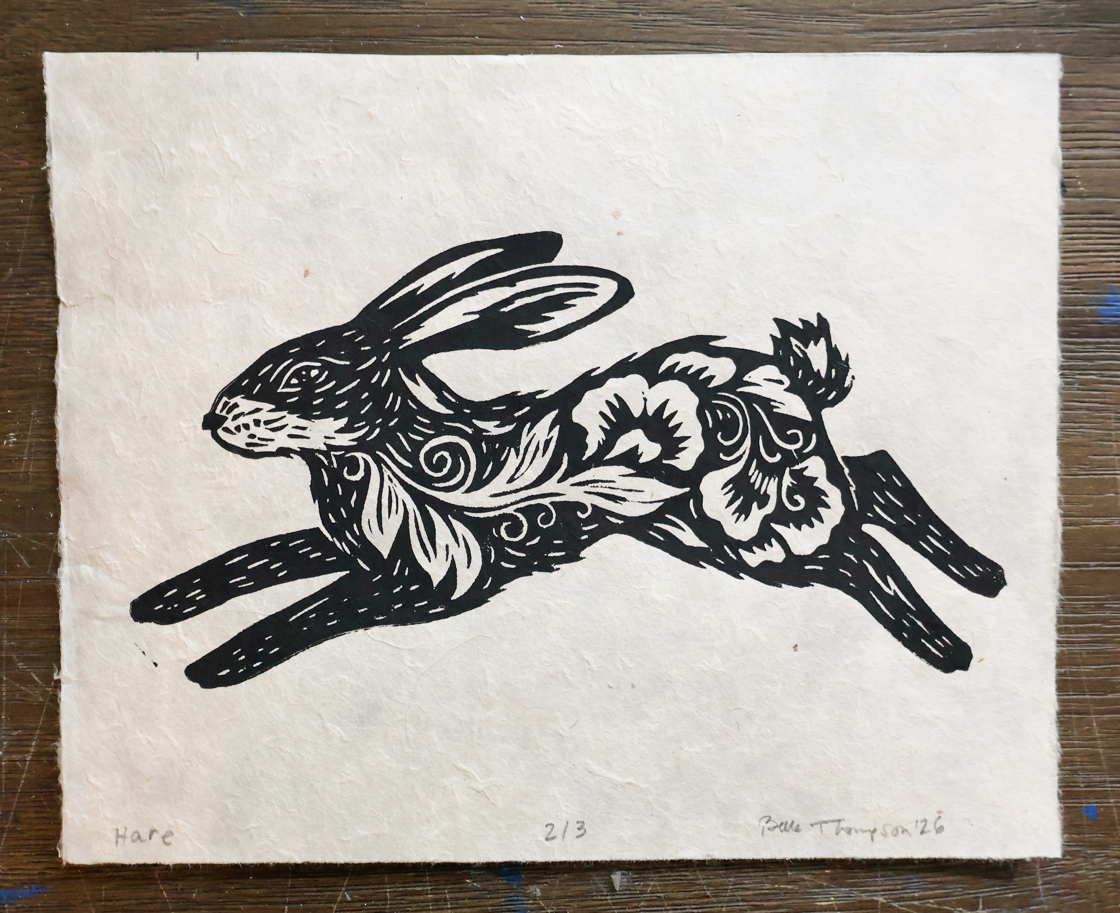 A square black-and-white art print on textured handmade paper, depicting a leaping hare with floral and swirl details, placed on a dark wood surface.