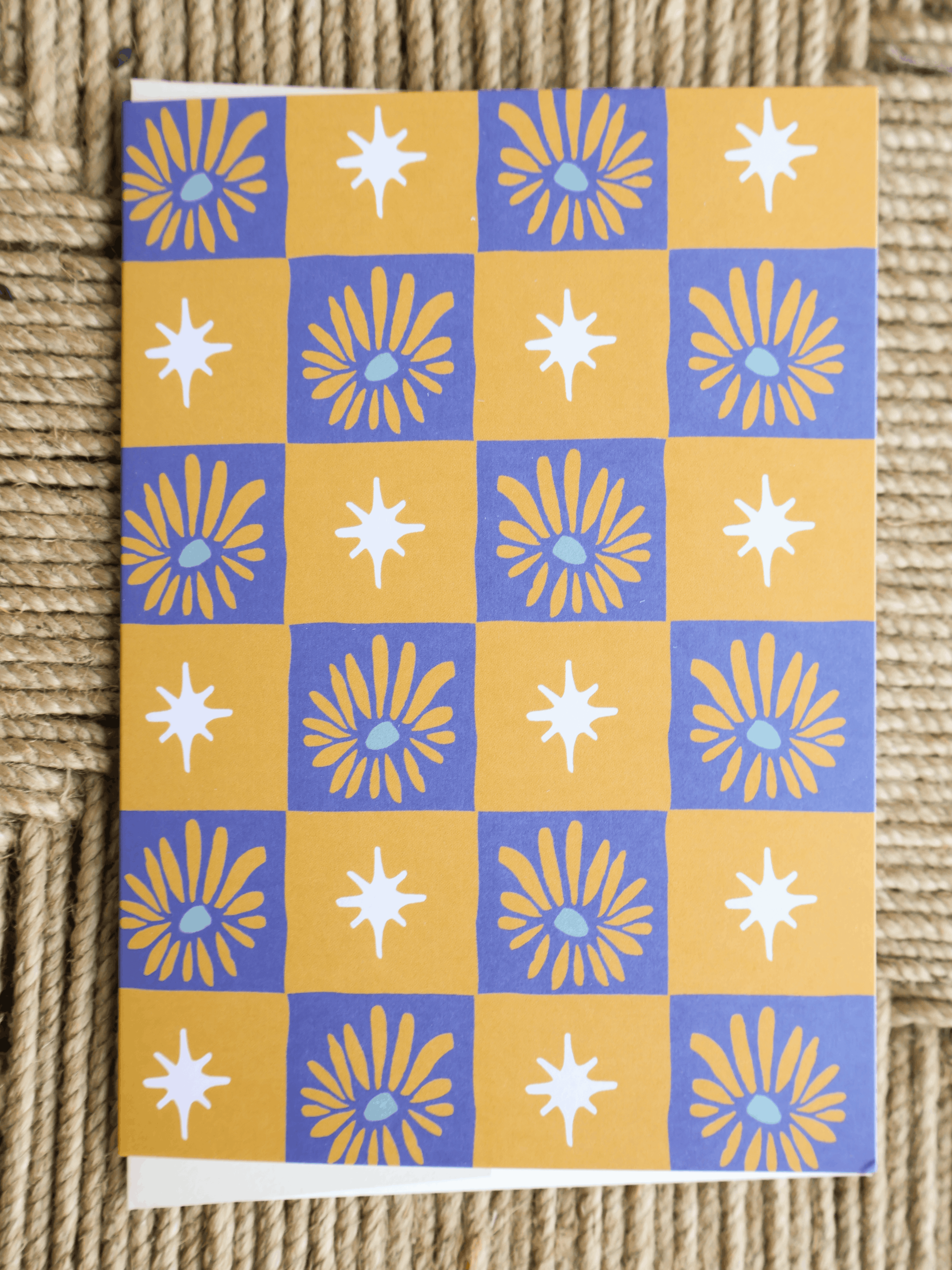 A square art print or card featuring a repeating checkerboard pattern of yellow and blue squares, each containing white starburst or sunburst motifs, photographed on a woven beige surface.