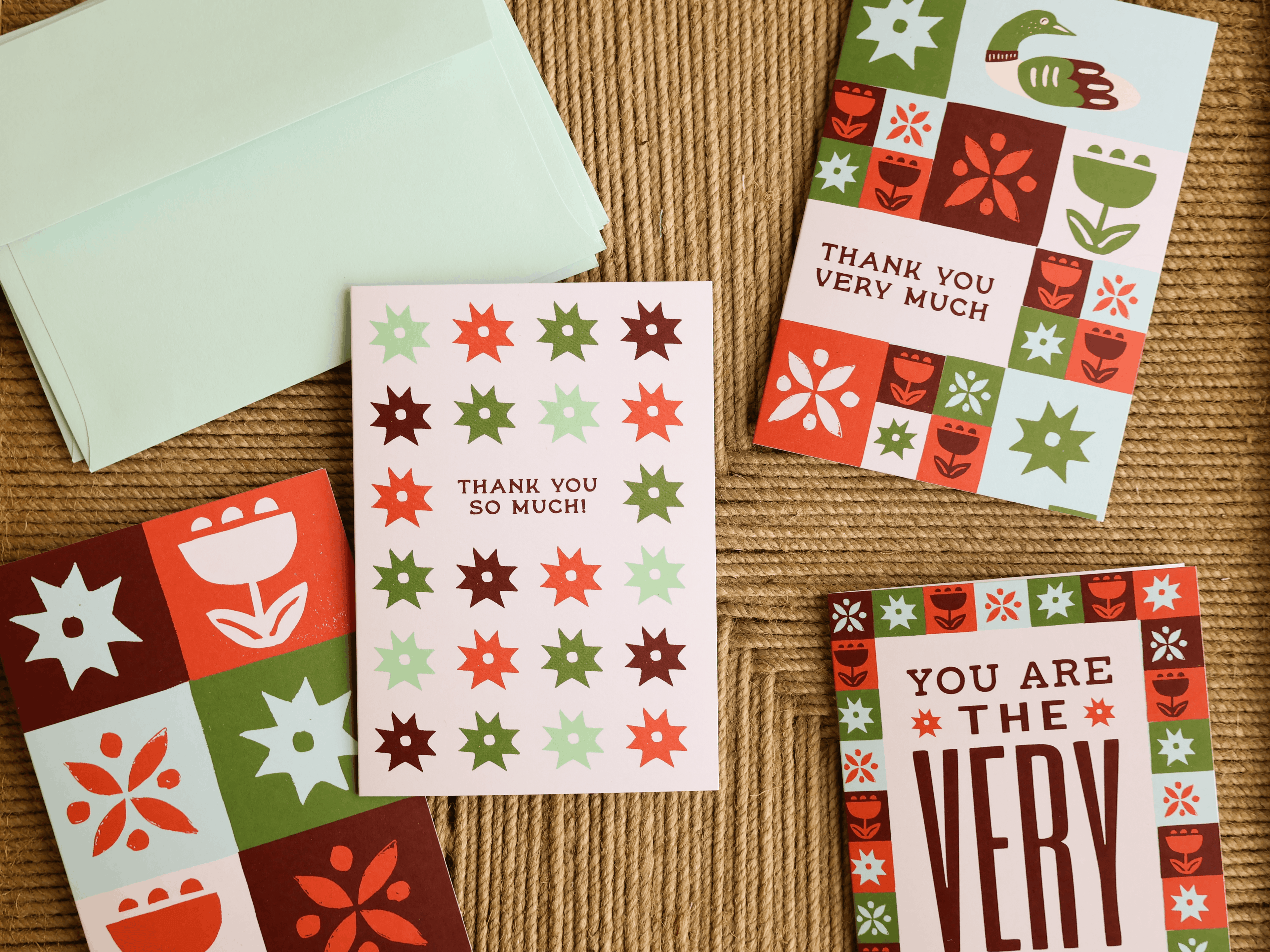 A set of thank you cards with retro floral and geometric patterns in red, green, and orange, displayed on a textured brown kraft paper background with a mint green envelope.
