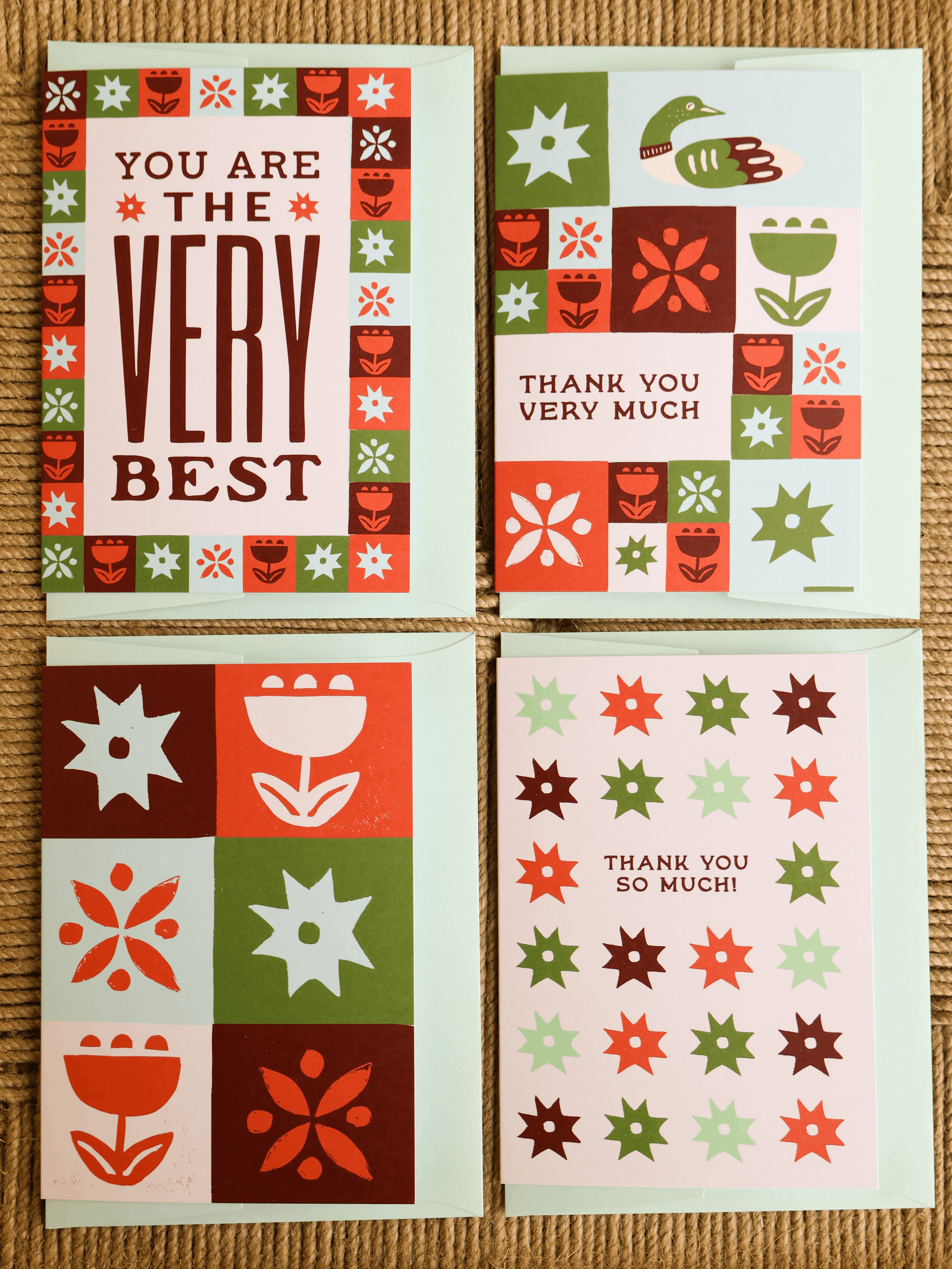 Four greeting cards arranged in a 2x2 grid on a woven beige surface. The cards feature retro patchwork floral and geometric designs in red, green, white, and dark brown, with messages including 'YOU ARE THE VERY BEST', 'THANK YOU VERY MUCH', and 'THANK YOU SO MUCH!'.