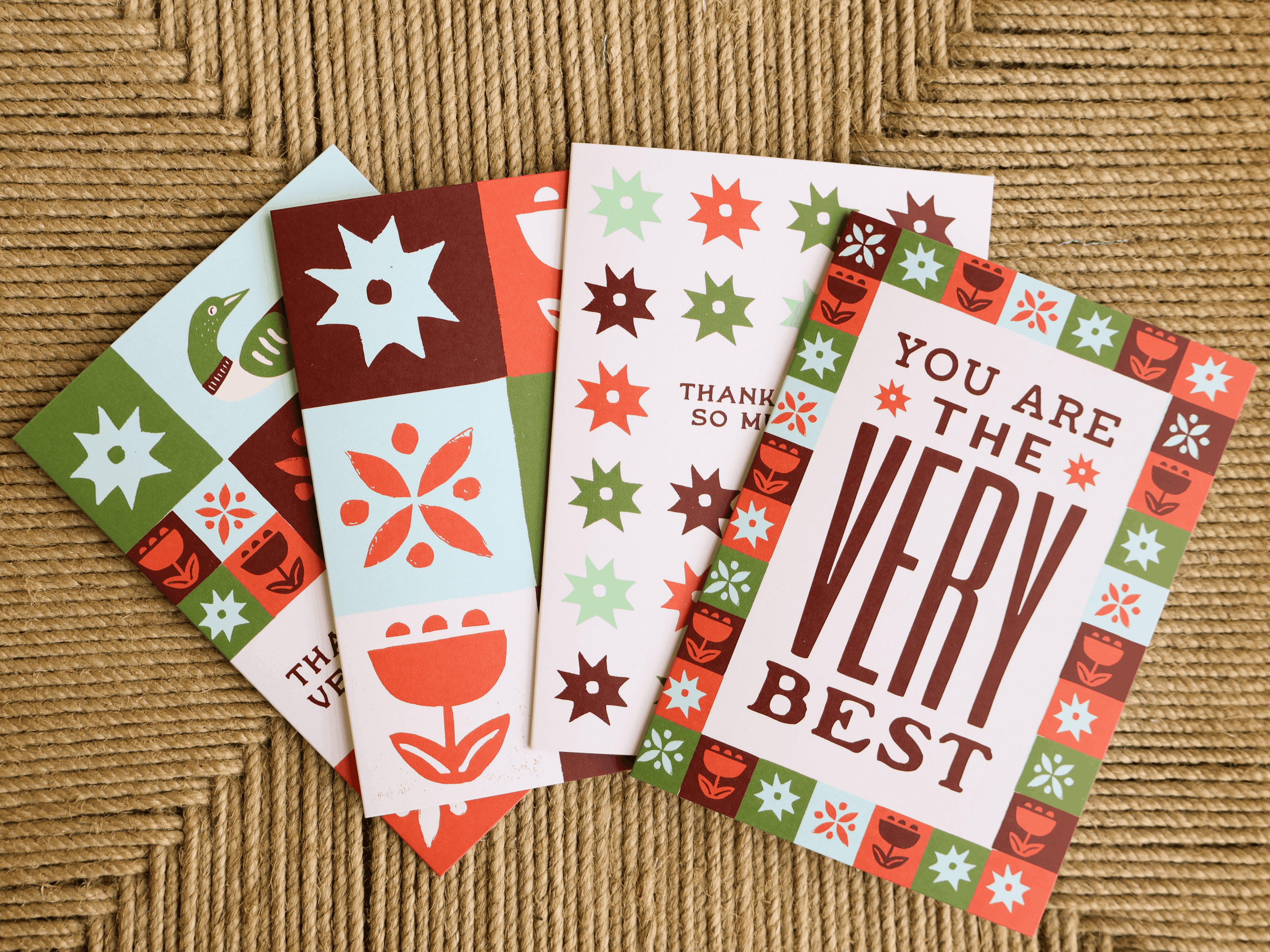 A set of five colorful patterned greeting cards fanned out on a woven straw surface. The cards feature retro geometric designs in red, orange, green, and brown, with one card reading 'YOU ARE THE VERY BEST' and another reading 'THANK SO MUCH'.