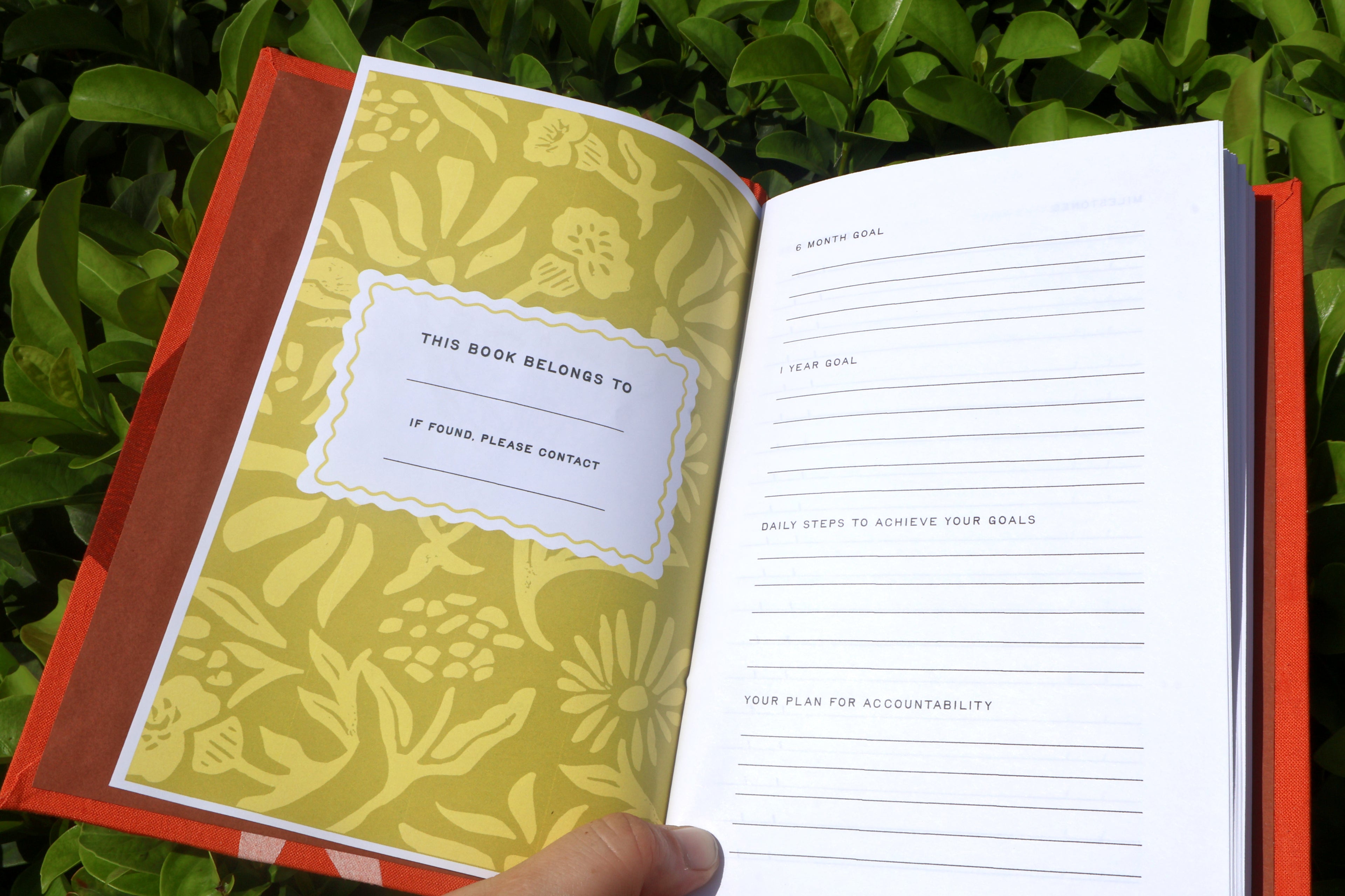 An open red hardcover self care reflections log held by a hand, showing a yellow floral-patterned cover page with a 'This book belongs to' label and white lined goal-setting pages inside, photographed outdoors against green foliage. hand bound and linocut printed with more love studio