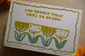 A square art print or card featuring the phrase 'YOU DECIDE WHAT GETS TO BLOOM' above a row of tulip-like flowers in yellow, green, and teal. The piece is photographed on a warm wood surface with partial orange objects at the edges.