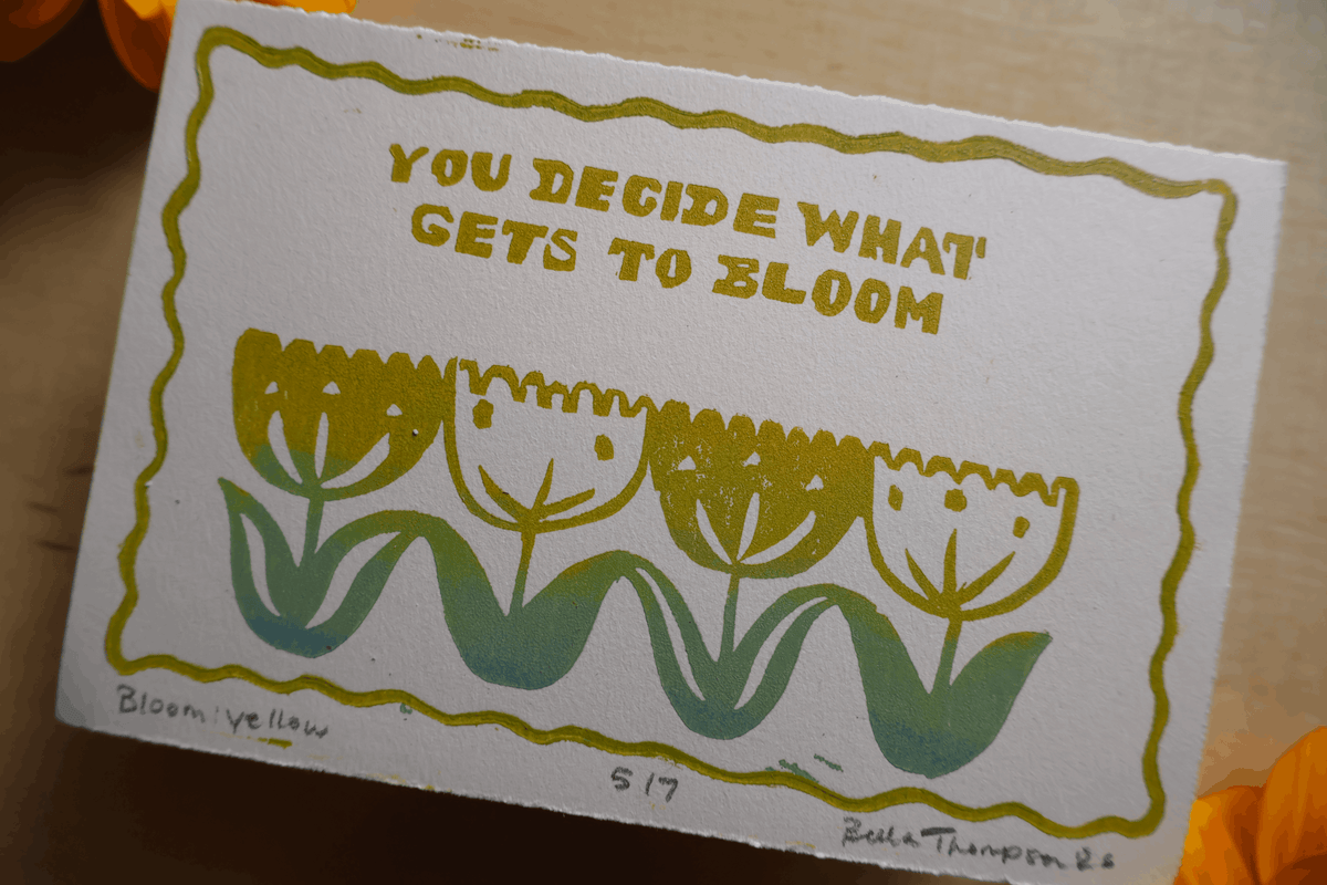 A square art print or card featuring the phrase 'YOU DECIDE WHAT GETS TO BLOOM' above a row of tulip-like flowers in yellow, green, and teal. The piece is photographed on a warm wood surface with partial orange objects at the edges.