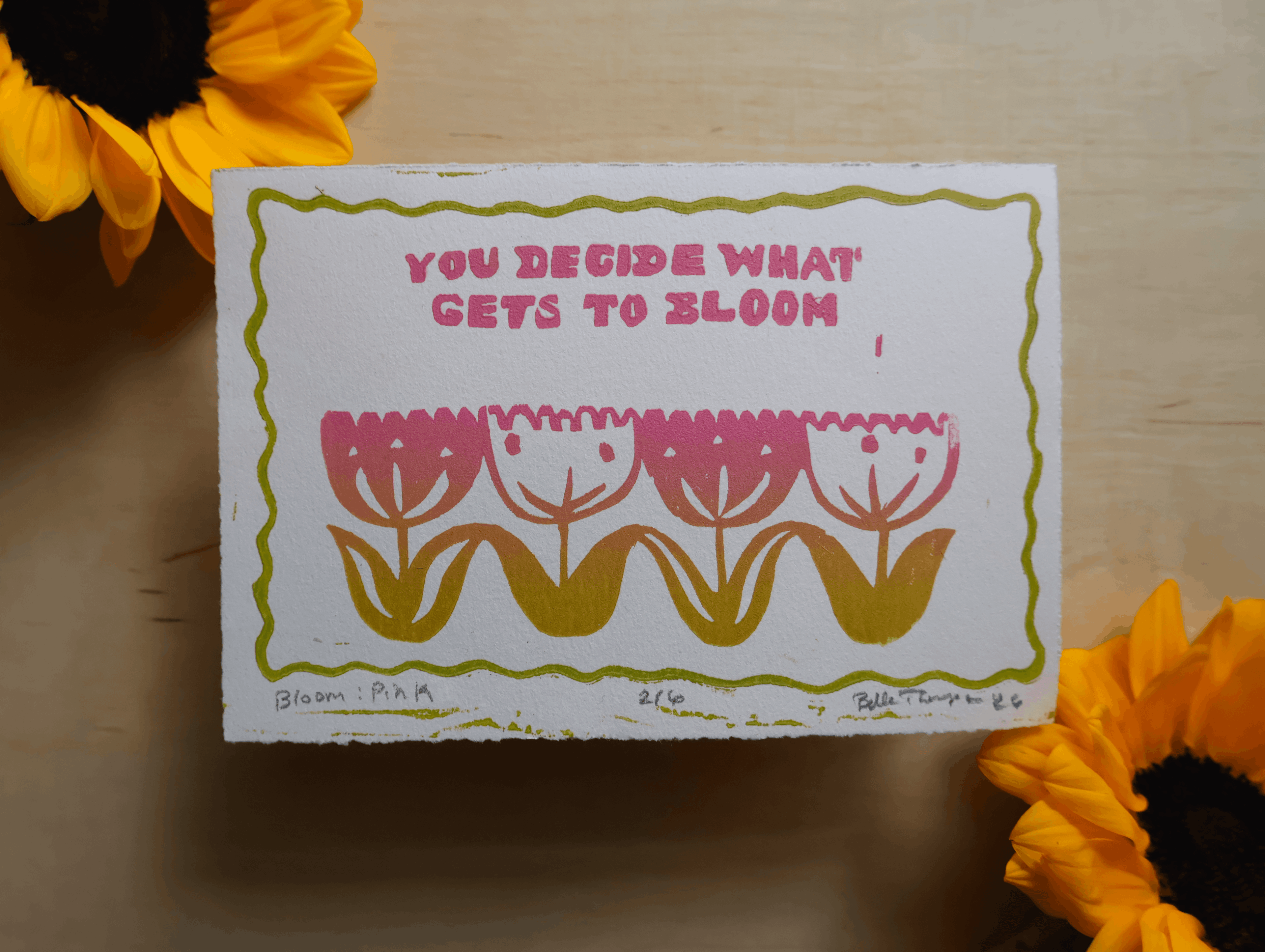 A square white art print or card with a hand-drawn olive green border, pink text reading 'YOU DECIDE WHAT GETS TO BLOOM', and stylized pink and mustard flowers, photographed on a light wood surface with sunflower petals at the corners.